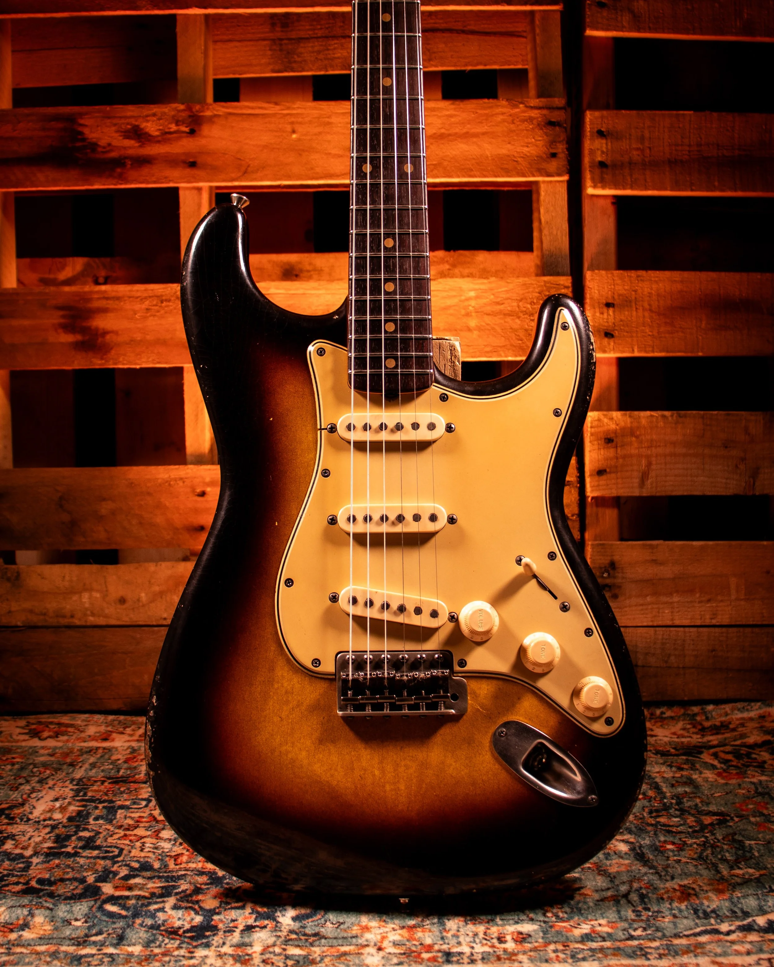 An electric guitar with a sunburst finish, white pickguard, and black body, resting on a wooden floor with a patterned rug in the background.