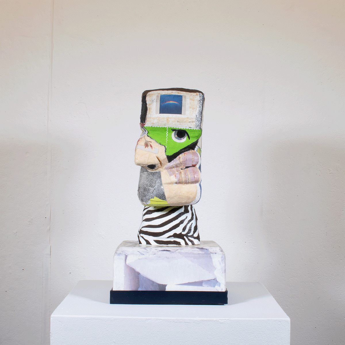 Mixed media sculpture of a face composed of various materials including fabric, paper, and paint, mounted on a pedestal in front of a plain wall.
