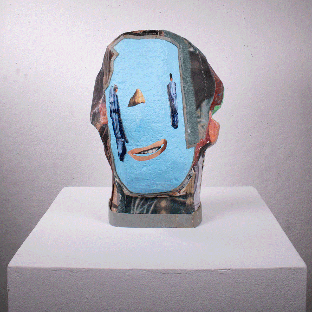 Mixed media art sculpture of a human head with a collage-style face, featuring a blue background, painted eyes with tears, and a smiling mouth, mounted on a white pedestal against a plain wall.