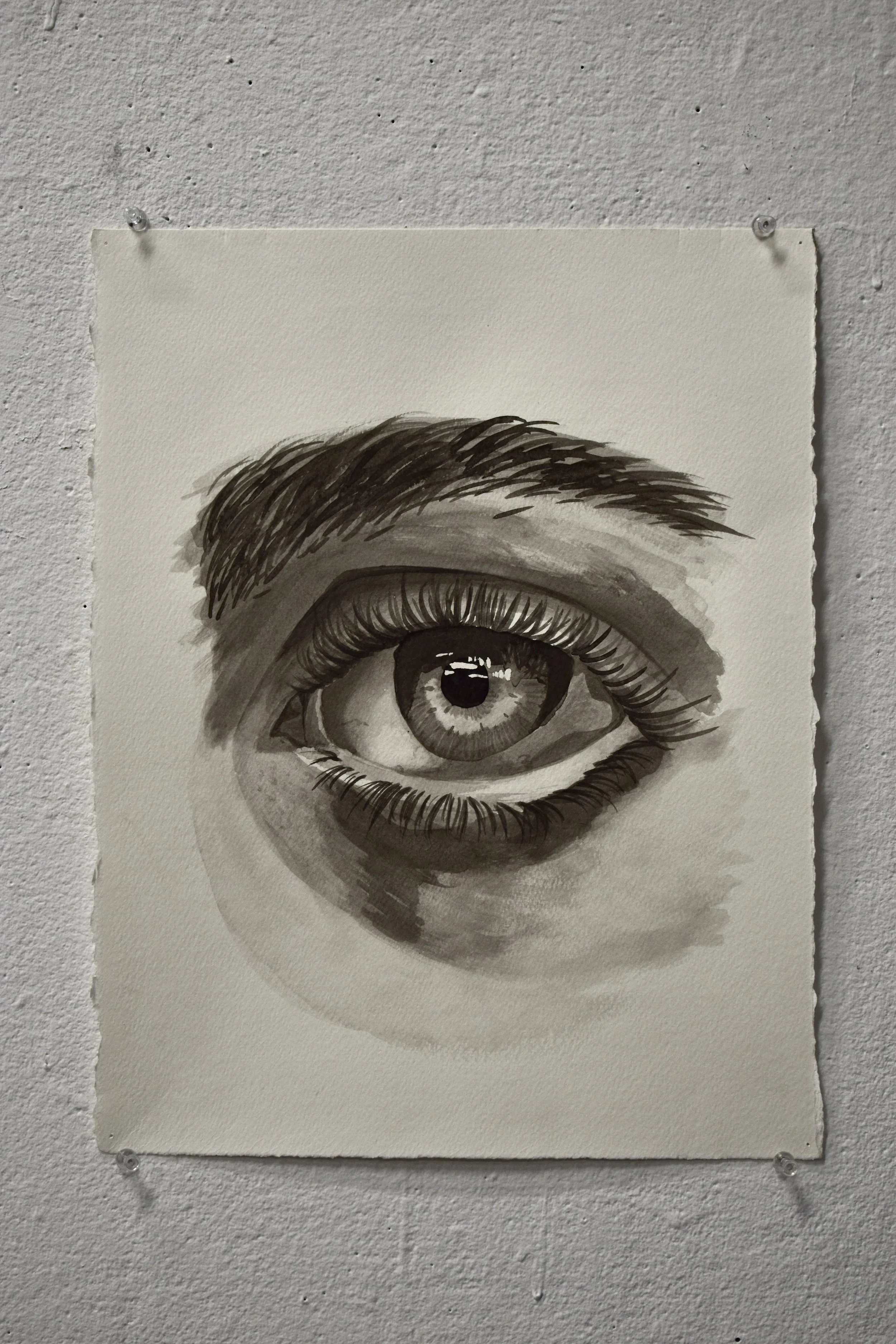 A black and white watercolor painting of a realistic human eye on textured paper pinned to a white wall.
