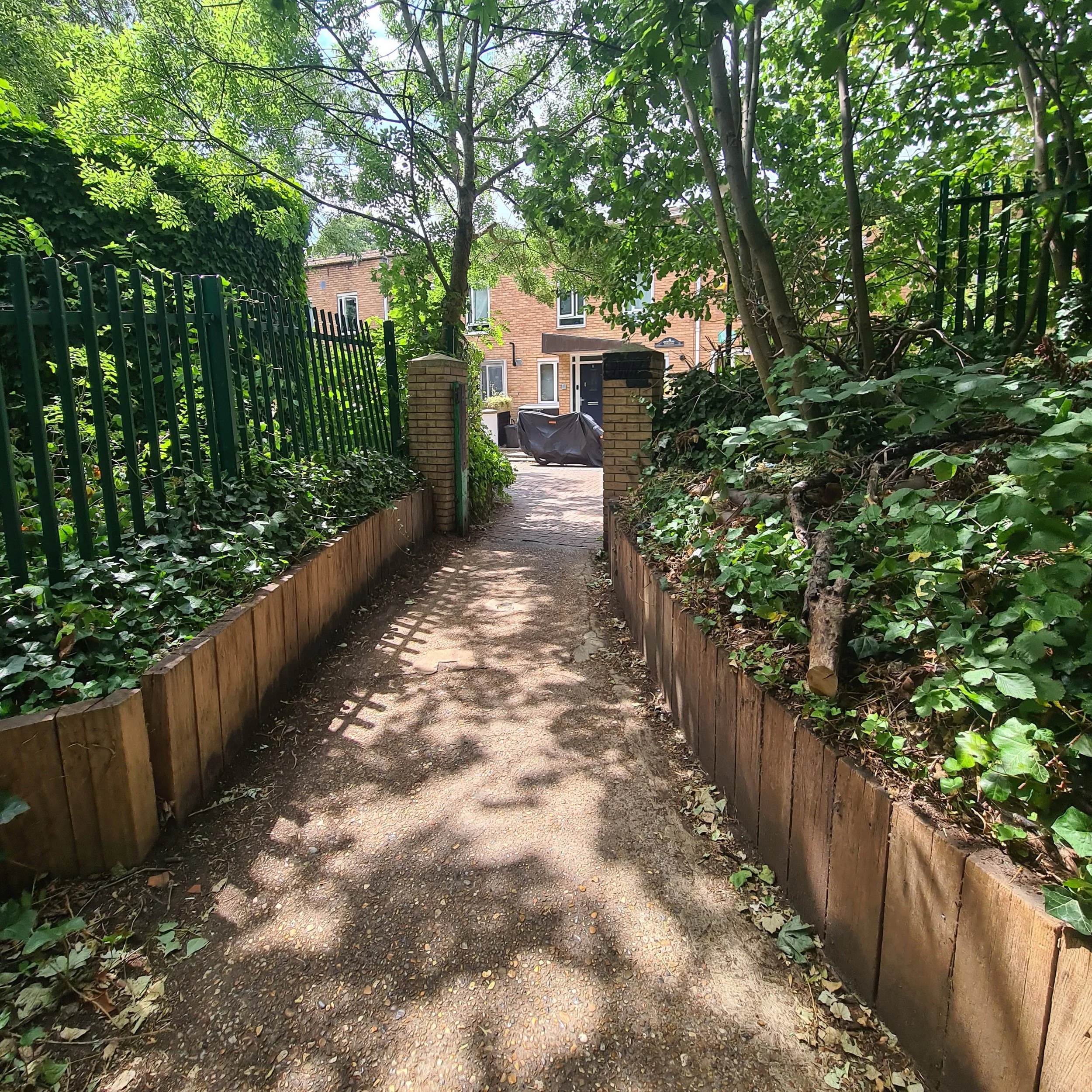 A narrow garden path flanked by low wooden planters filled with green ivy plants, leading to a brick paved area with brick pillars and a metal gate, surrounded by trees and leafy bushes.