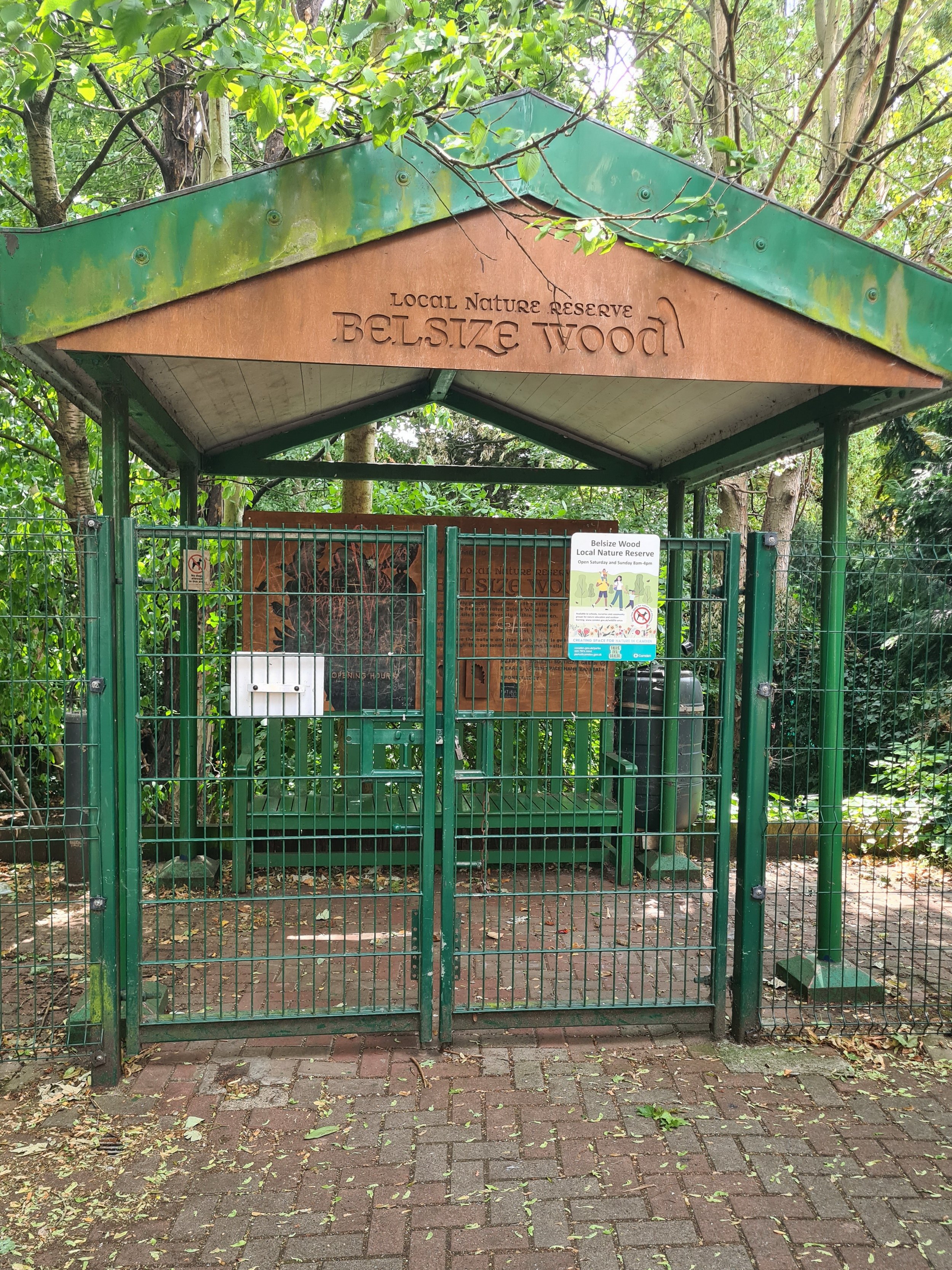 Gated enclosure at Belsize Wood Local Nature Reserve with wooden sign and information board surrounded by trees and greenery.