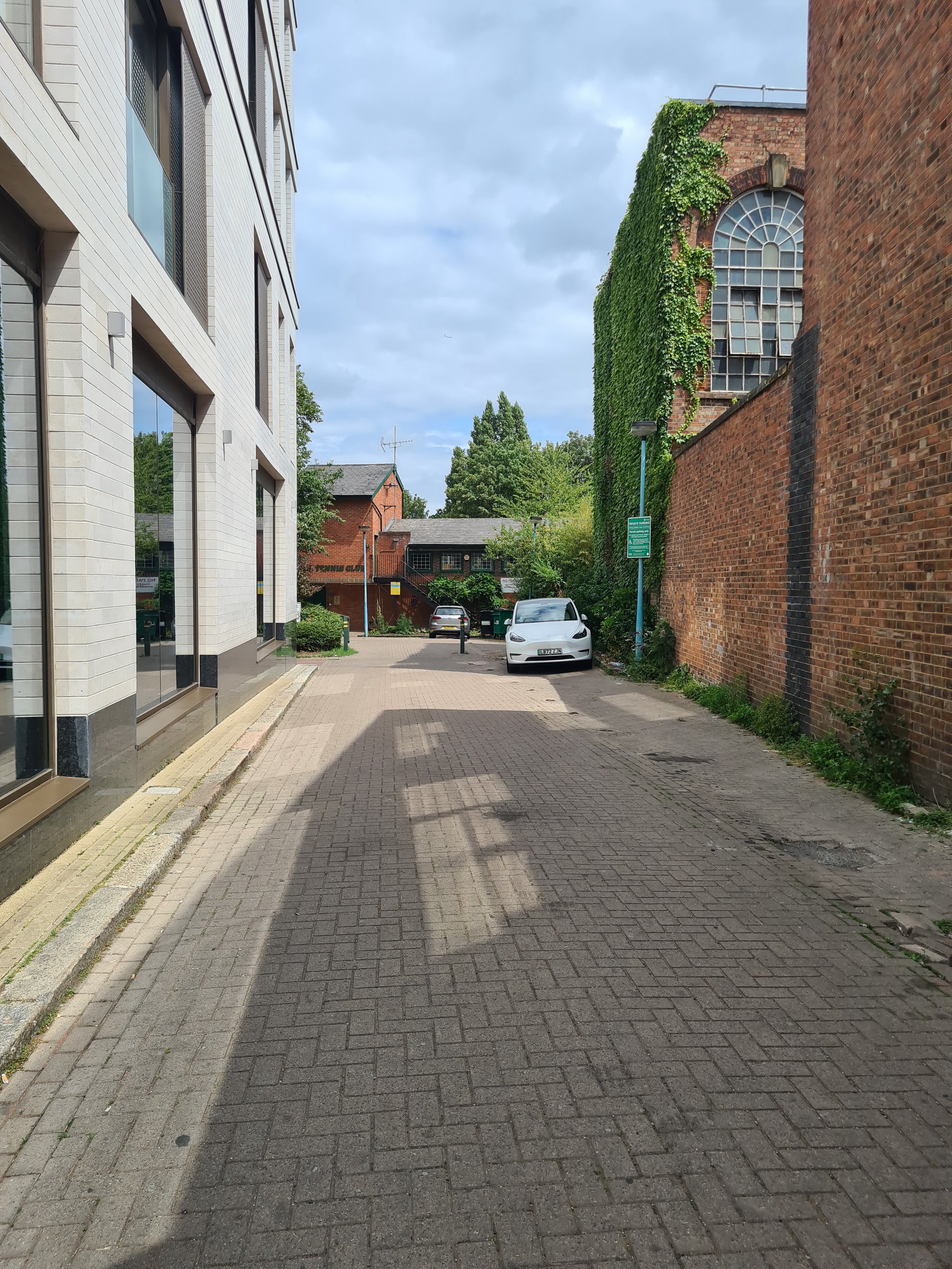 A narrow brick-paved alleyway between a modern white building on the left and an old brick building on the right covered in green vines. There is greenery and trees in the background under a partly cloudy sky.