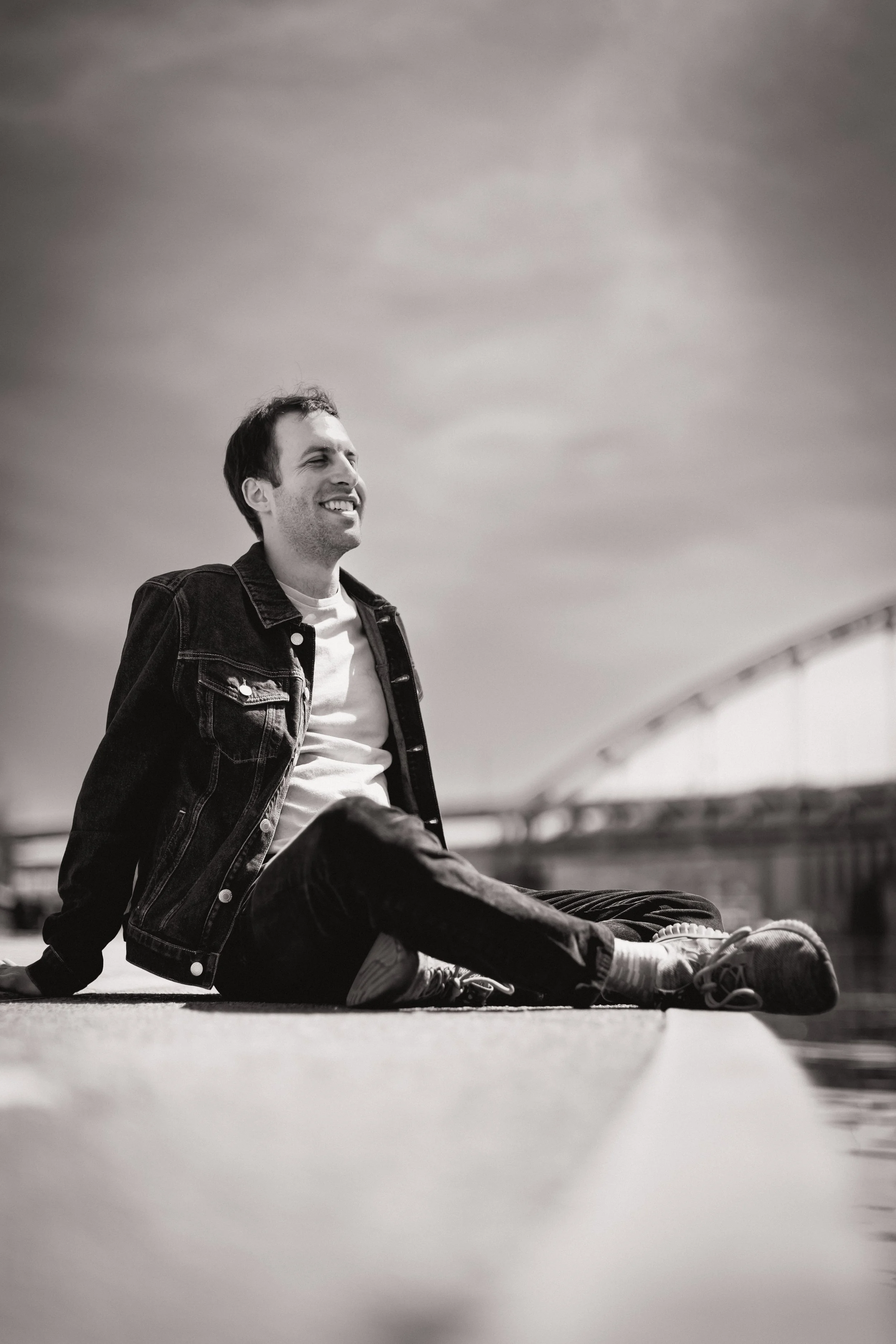 A young man with dark hair and light skin, wearing a denim jacket, white t-shirt, sneakers, and jeans, sitting on the ground outdoors, smiling and looking to the side with a cloudy sky above.
