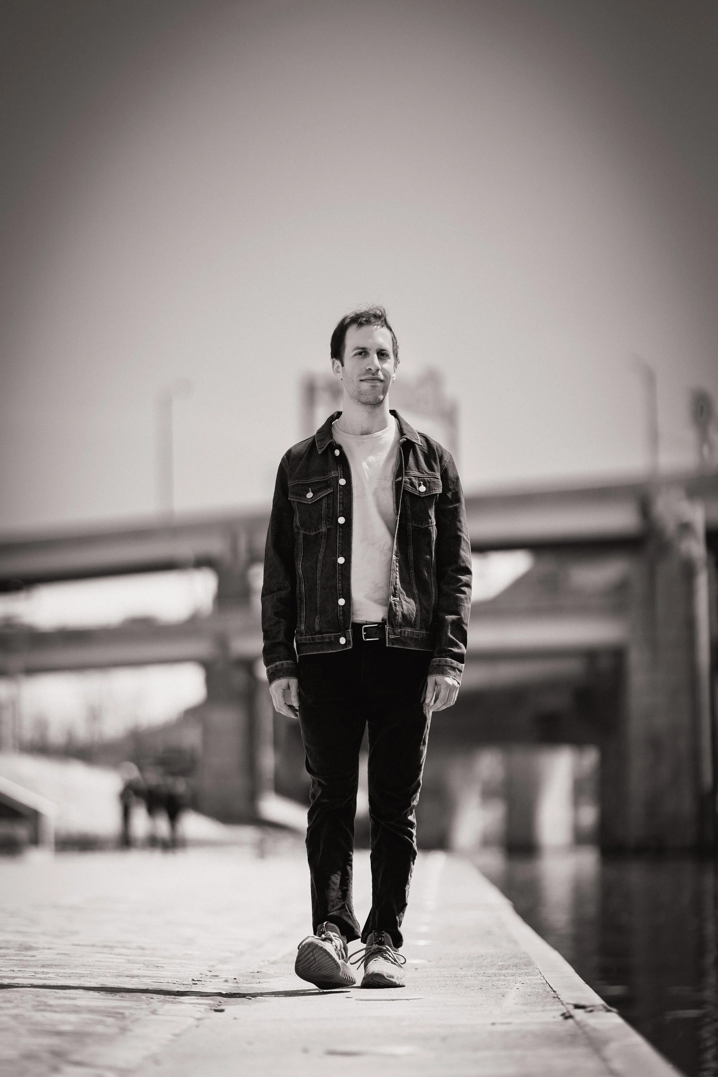 A young man with dark hair walks on a bridge or walkway near water, wearing a denim jacket, white t-shirt, black pants, and sneakers, with a background of an overpass and city skyline in black and white.