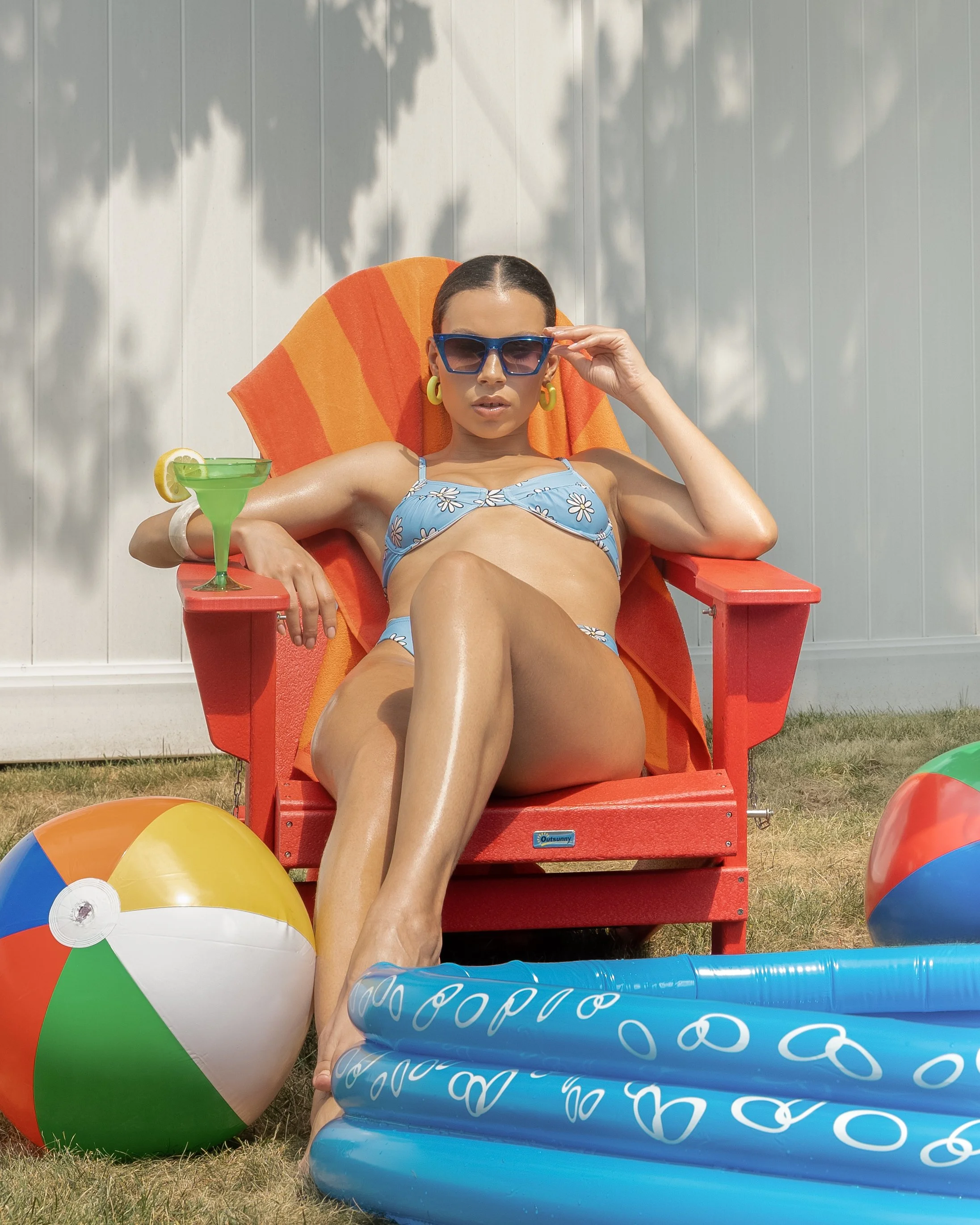 Woman in blue bikini sitting on a red chair with sunglasses and a drink, surrounded by beach balls and an inflatable pool.
