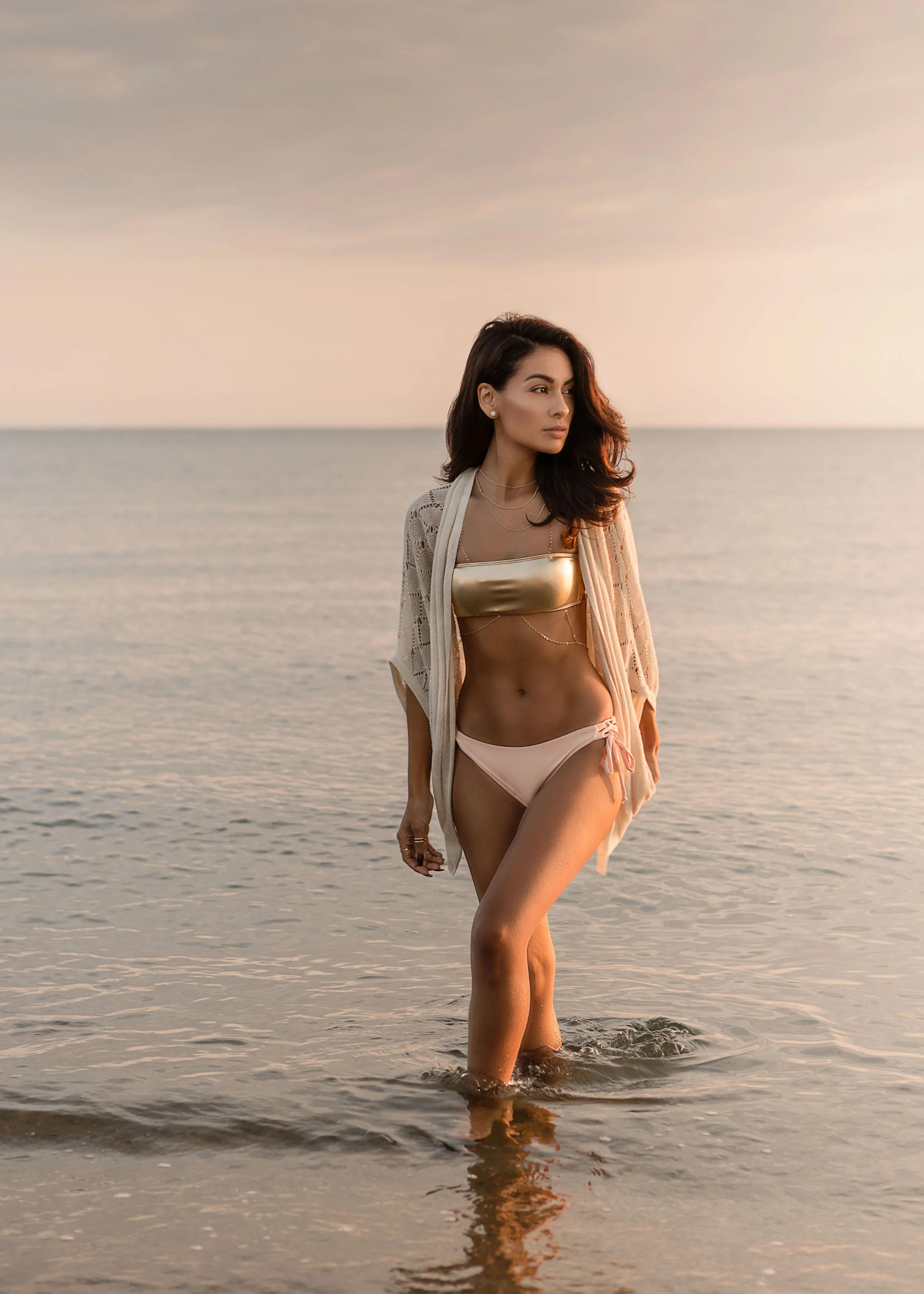 Woman in a gold bikini top and pink bottoms, standing at the beach with a light cover-up, during sunset.