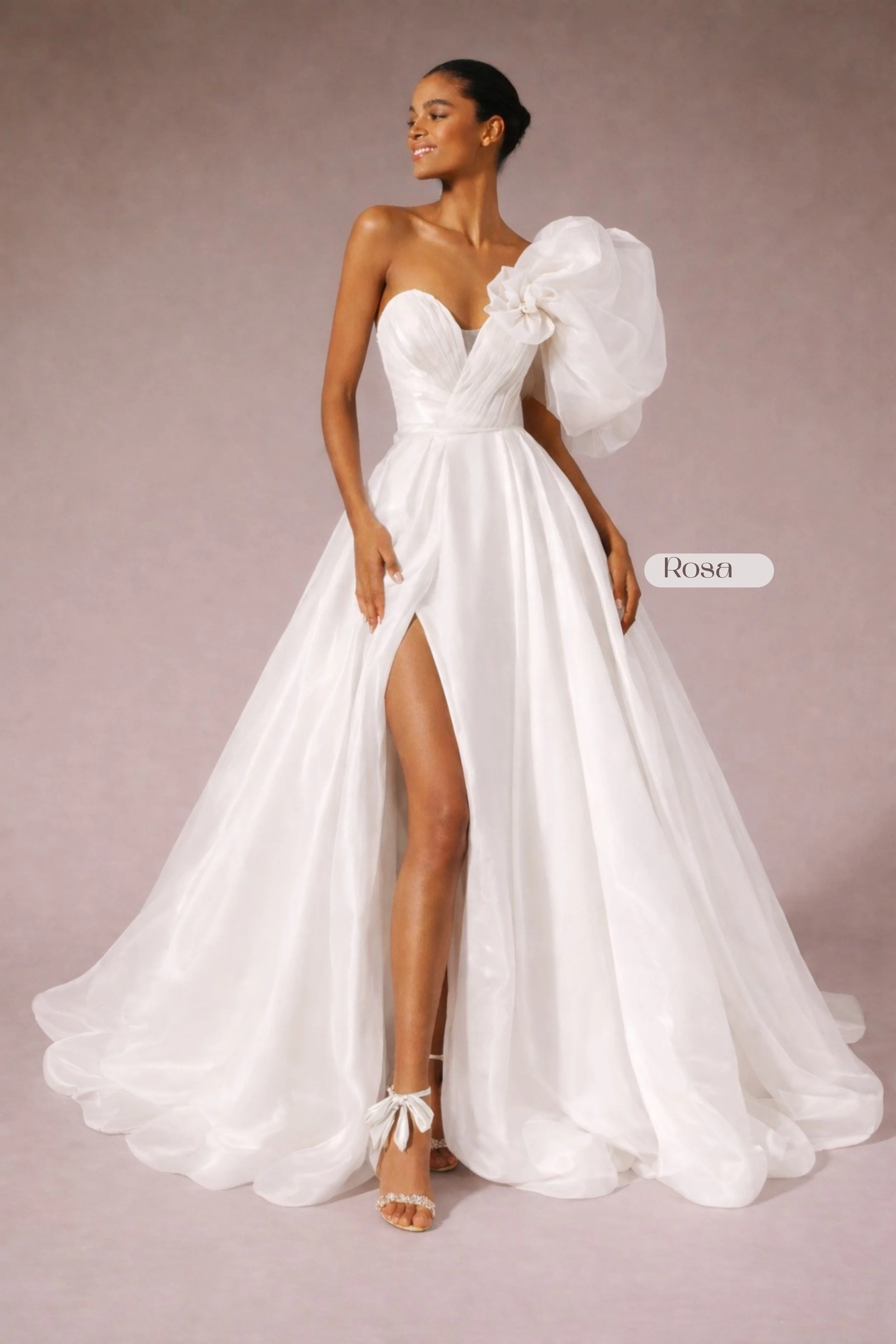 White one-shoulder wedding dress with voluminous organza sleeve, floral accent, pleated corset bodice, full skirt, high leg slit, and modern couture silhouette.