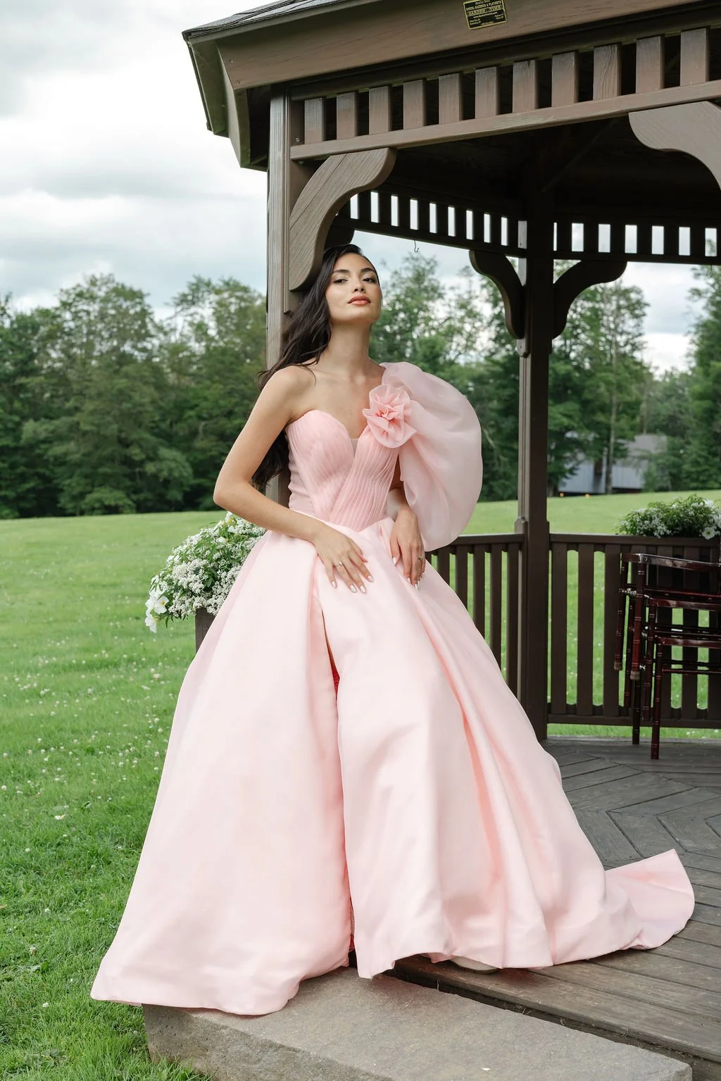 Blush pink one-shoulder wedding dress with voluminous organza sleeve, floral accent, pleated corset bodice, full skirt, high leg slit, and modern couture silhouette.