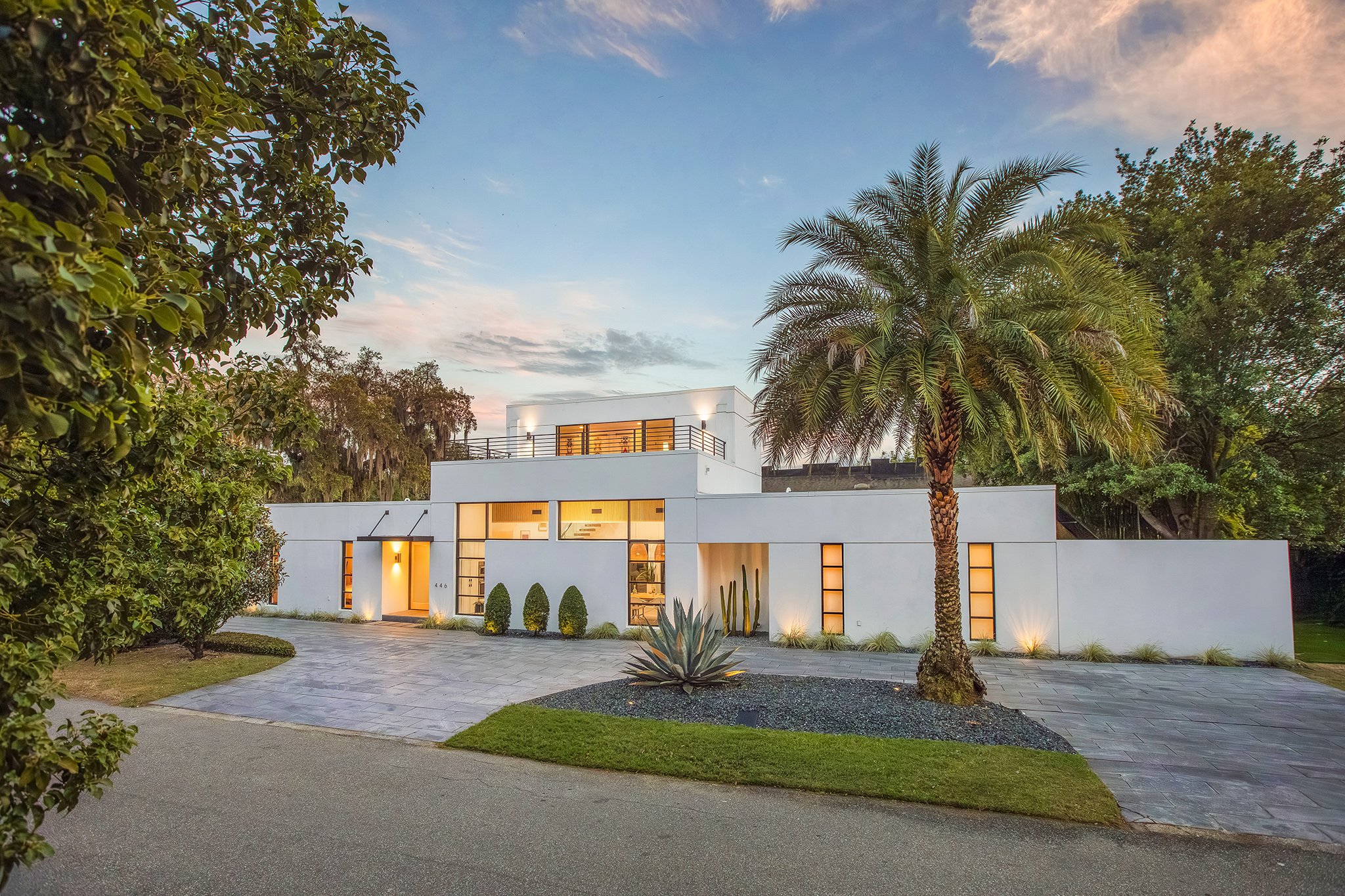 Modern white house with large glass windows and doors, palm trees, and landscaped front yard, under a partly cloudy sky at dusk.