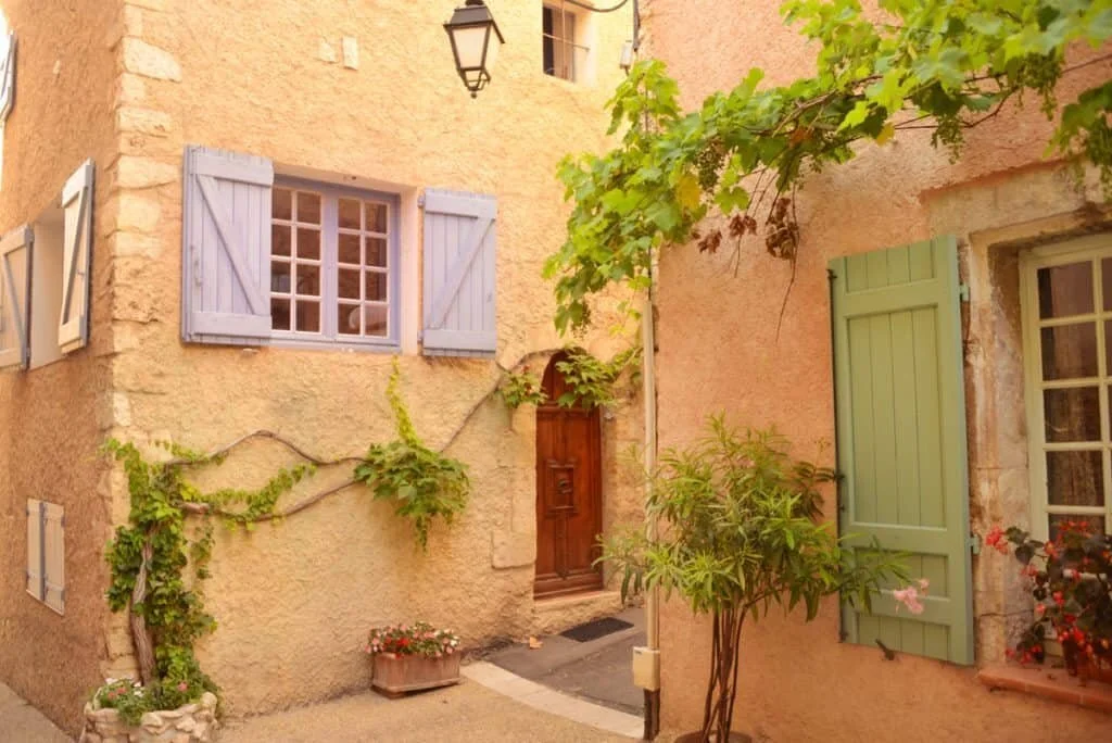 An illustration of a cozy European-style courtyard with pastel-colored walls, small windows with shutters, and lush green plants and flowers.