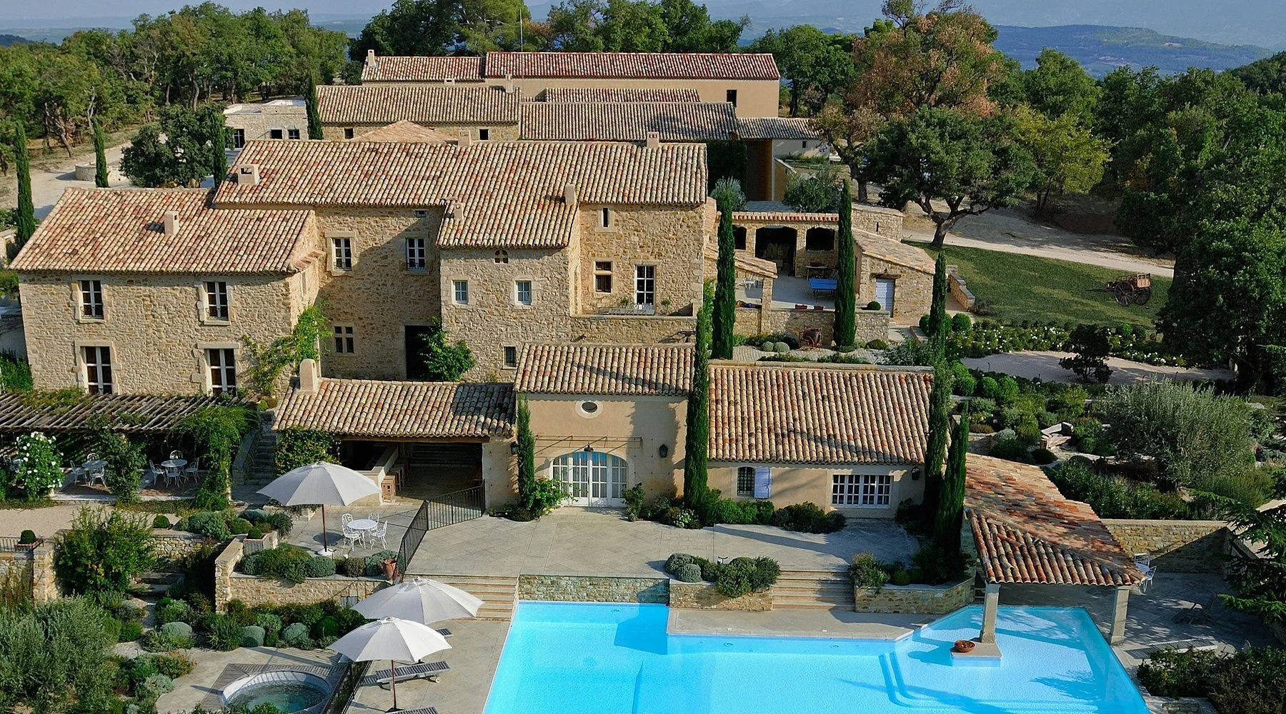 Aerial view of a Mediterranean-style villa with beige stone walls, red-tiled roofs, surrounded by lush green trees, a garden, and a swimming pool with lounge chairs and umbrellas.