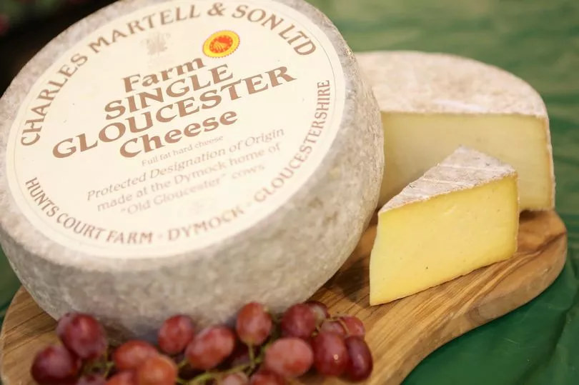 A wheel of Charles Martell & Son Ltd farm single Gloucester cheese on a wooden board, with a slice cut to reveal its yellow interior, accompanied by a bunch of red grapes, all set on a green surface.