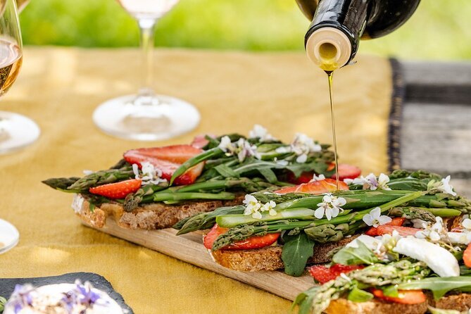 A wooden board with slices of bread topped with asparagus, strawberries, and edible flowers, with a bottle of olive oil pouring over it, and two glasses of white wine in the background.