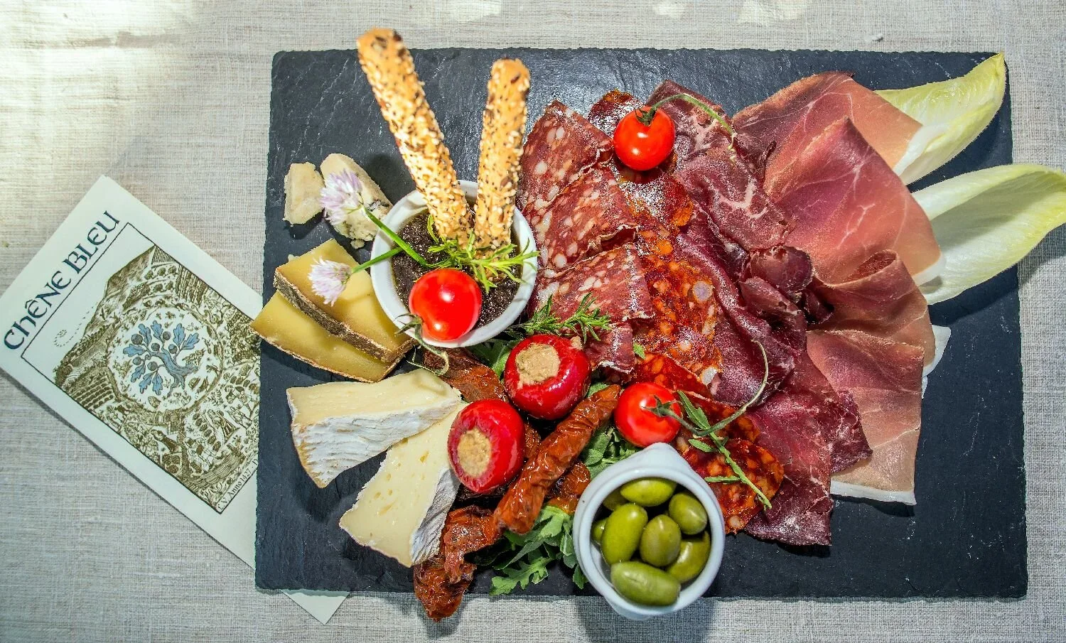 A cheese and charcuterie board with various cheeses, cured meats, cherry tomatoes, green olives, breadsticks, and garnishes, served on a black slate platter. An Italian newspaper is partially visible on the side.