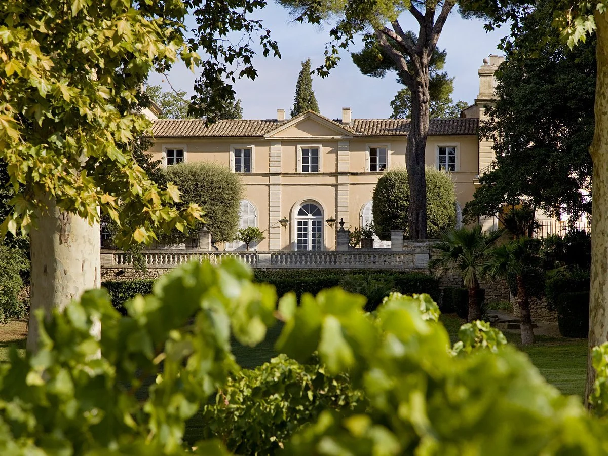 A large, classic style mansion surrounded by lush green trees and bushes.