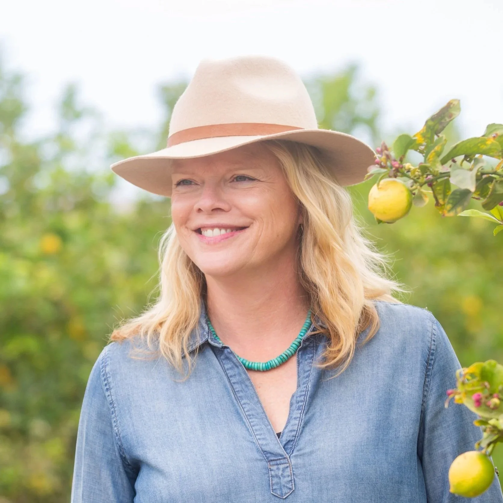 A woman smiling outdoors in an apple orchard wearing a wide-brimmed hat, a denim shirt, and a turquoise necklace.