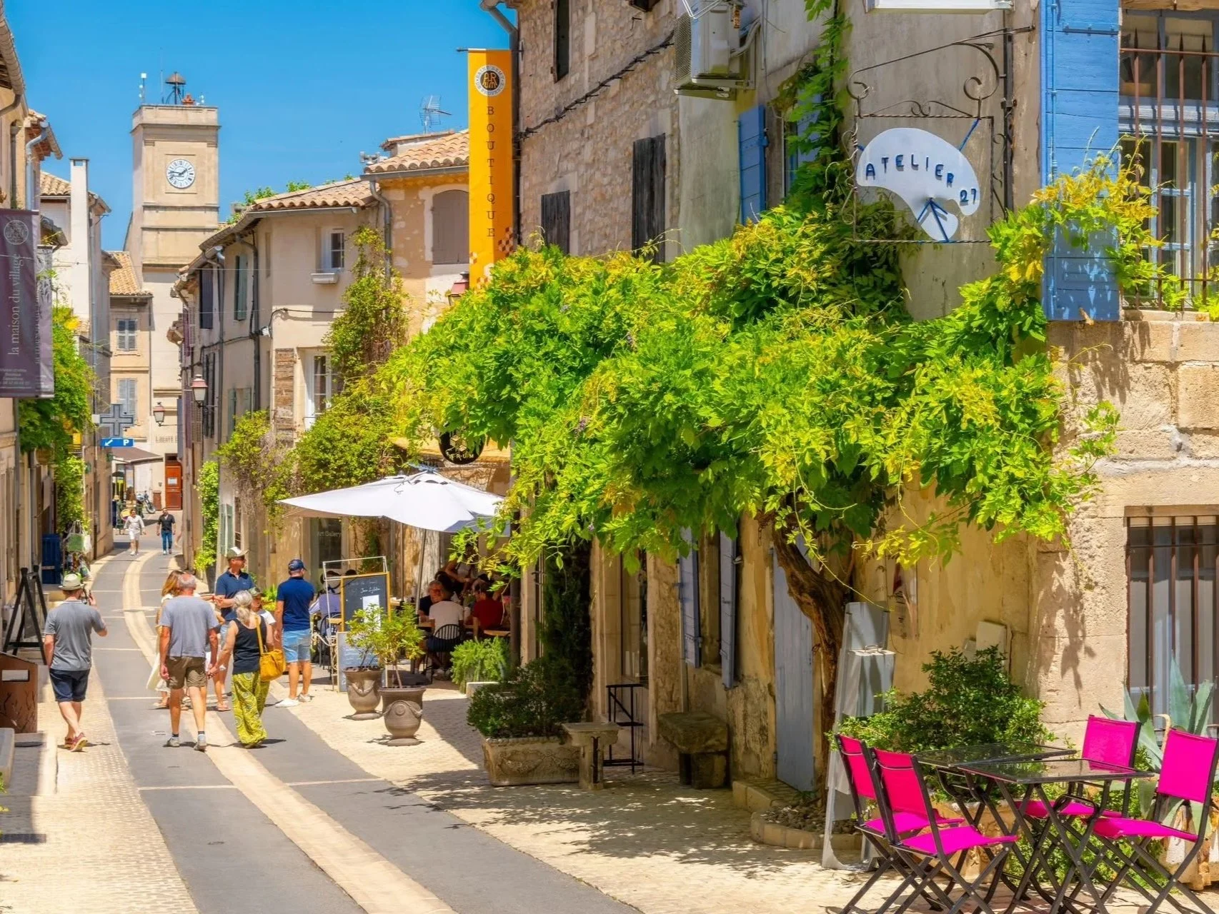 A sunny street scene in a European town with pedestrians, outdoor seating, greenery, and colorful buildings.