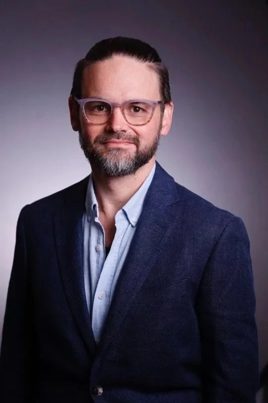 A man with glasses, a beard, and neatly styled dark hair, wearing a dark blue blazer and a light blue collared shirt, posing against a gradient dark background.