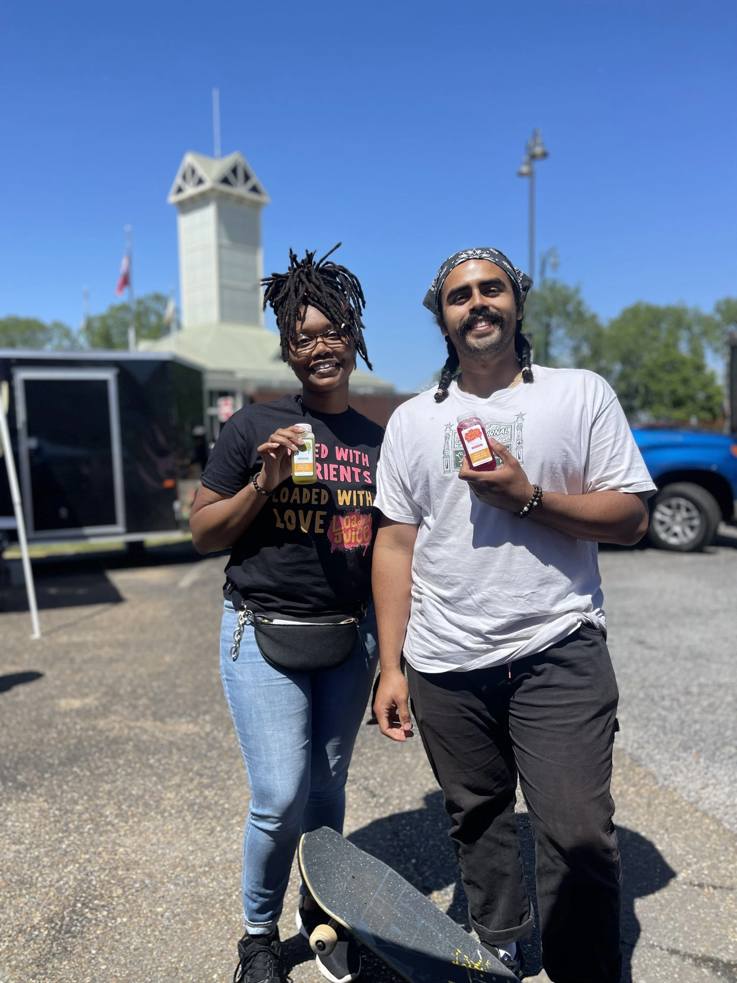 Two people smiling with juice bottles, one holding a skateboard, standing outdoors with a sunny sky and a tower in the background.