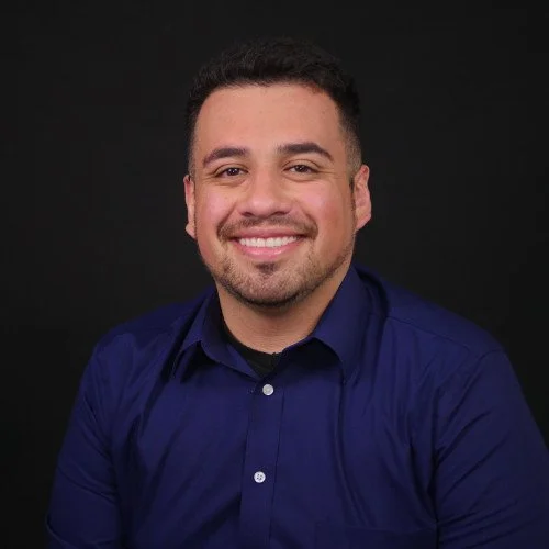 Portrait of a smiling man with short curly hair, wearing a dark blue button-up shirt, against a black background.