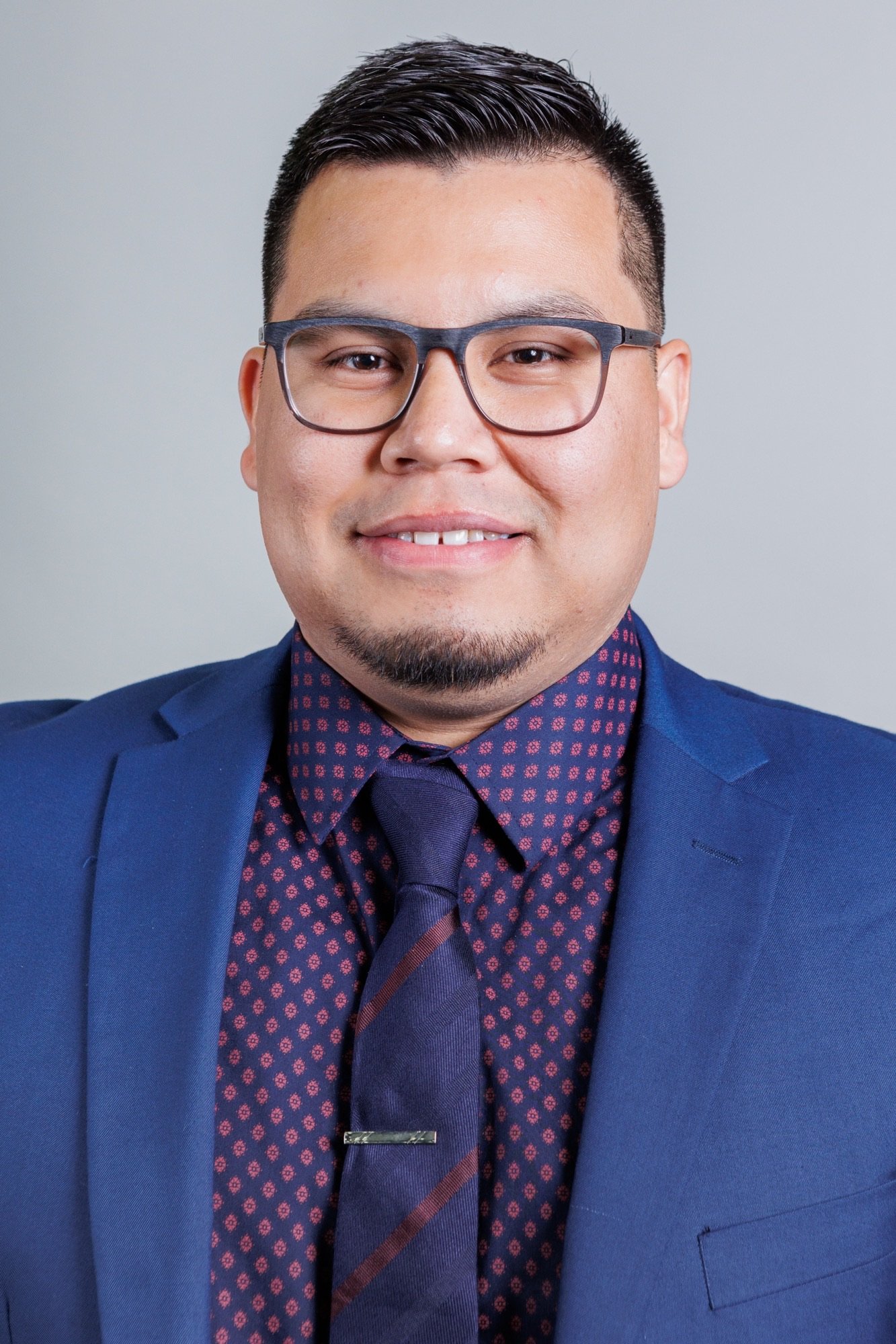 A man with glasses is wearing a blue suit, a dark patterned shirt, and a matching tie, smiling at the camera against a plain gray background.