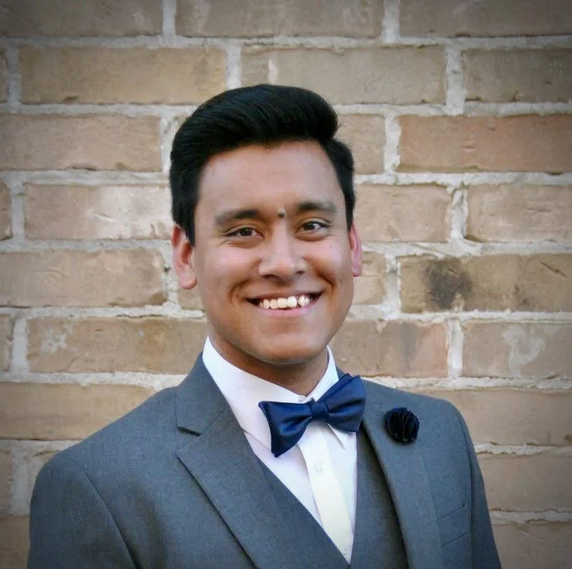 A man smiling, wearing a gray suit with a blue bow tie, standing in front of a brick wall.