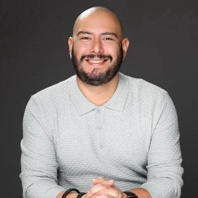 A smiling man with a beard and shaved head in a light gray collared shirt against a dark background.