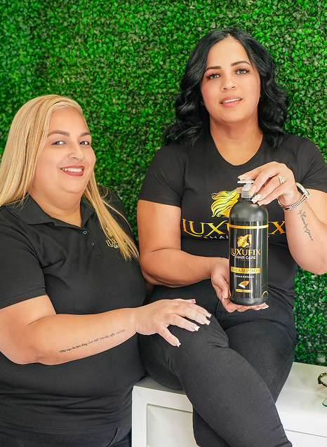 Two women in black shirts sitting in front of a green foliage wall, one holding a bottle of LUXE hair care product.