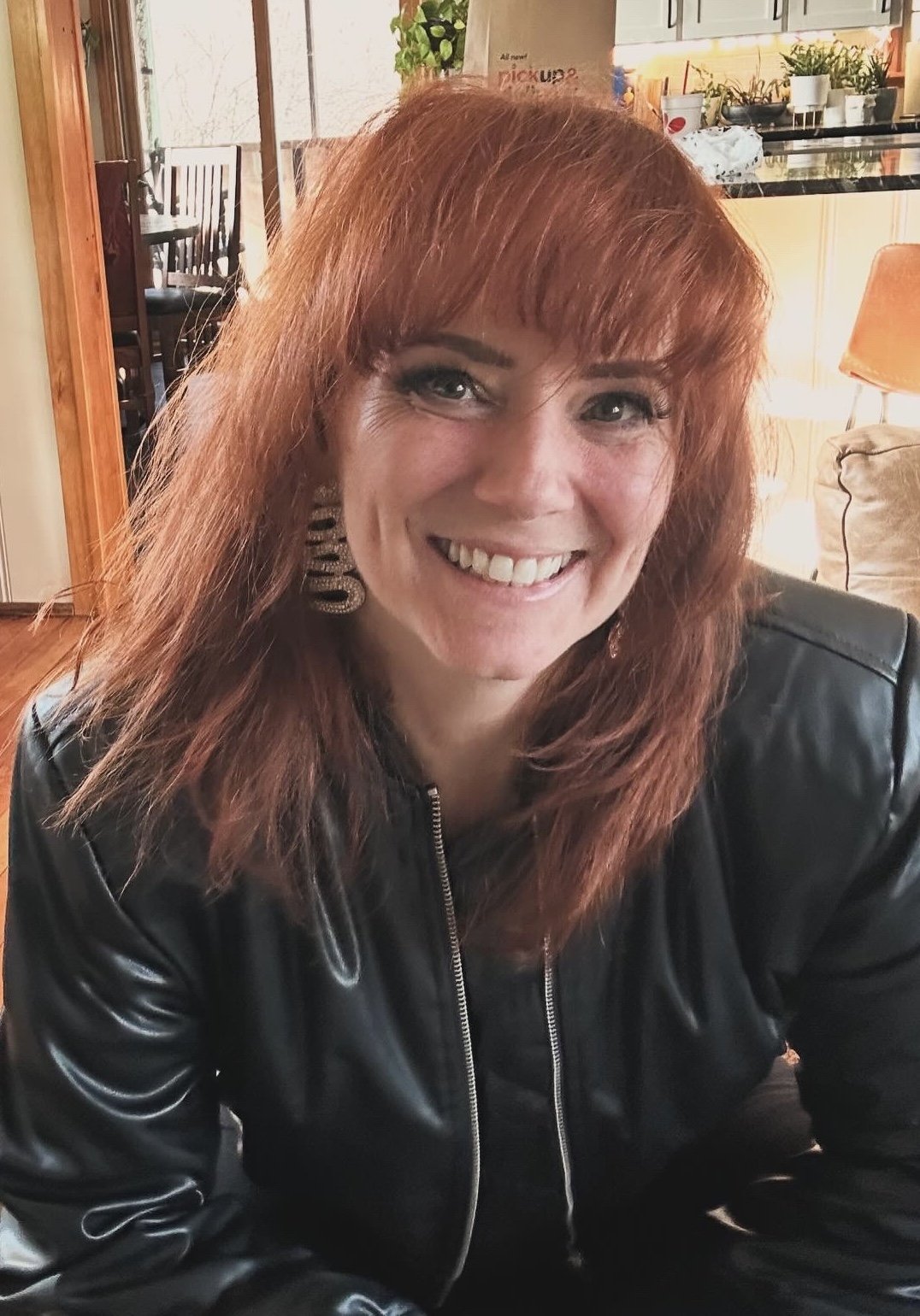 A woman with red hair, wearing a black leather jacket, smiling in a cozy home setting with plants and kitchen in the background.