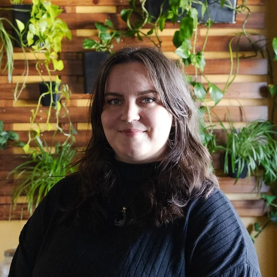 A woman with wavy brown hair, wearing a black top, smiling in front of a wooden wall with hanging green plants.
