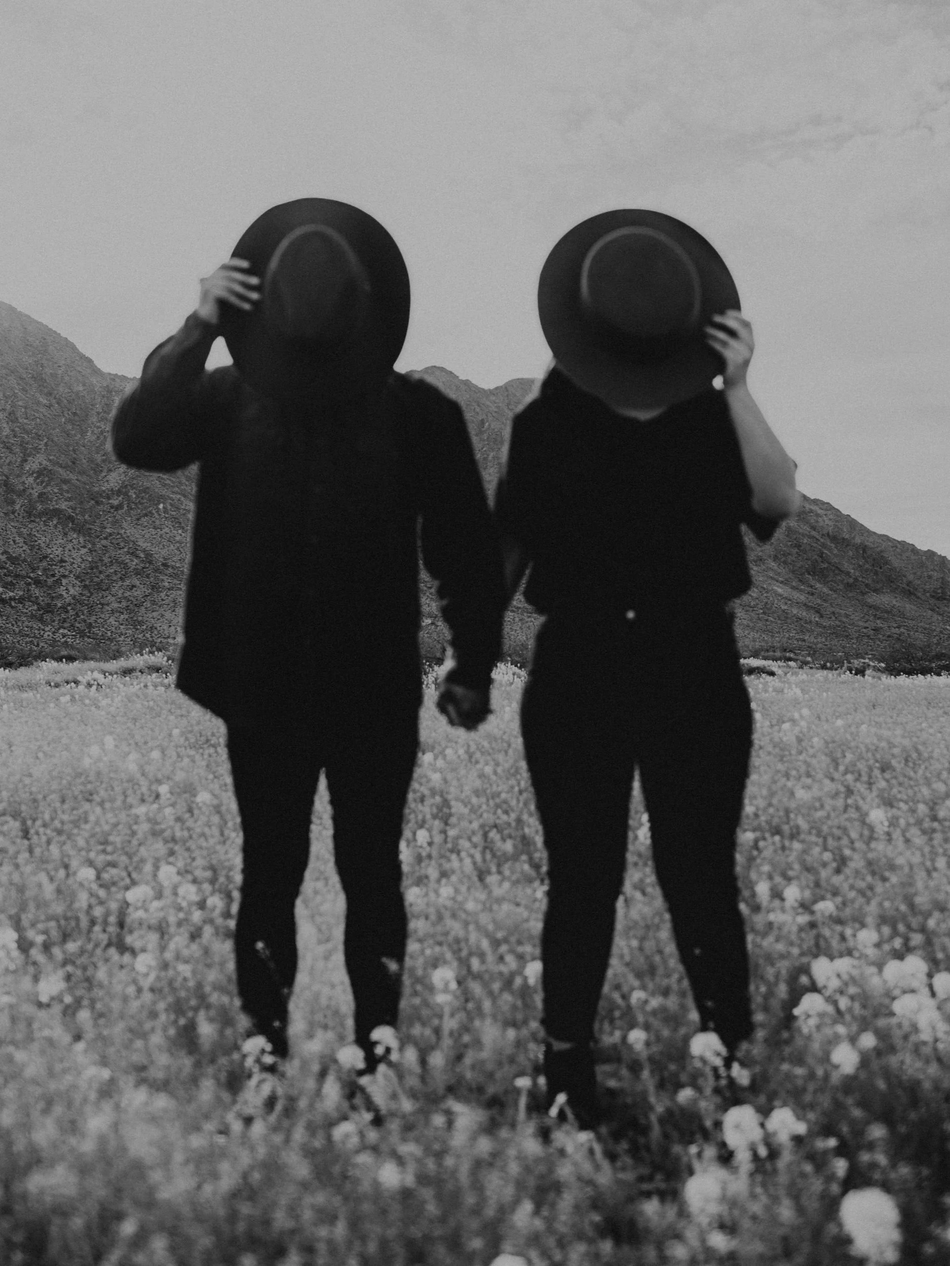 Two people walking hand in hand through a field of flowers, each holding a wide-brimmed hat in front of their faces, with mountains and sky in the background.
