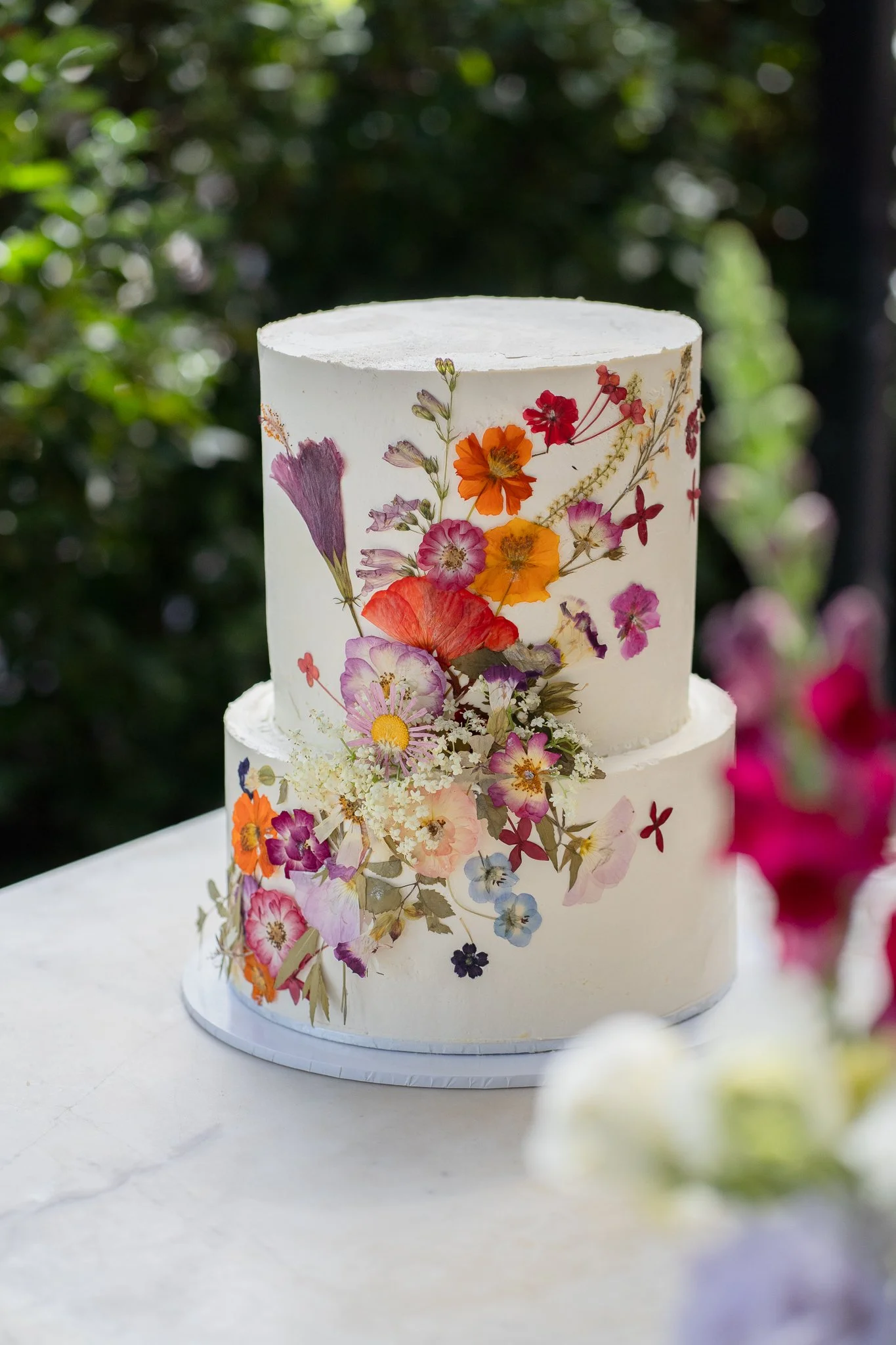 A white, two-tiered cake decorated with colorful pressed flowers on the sides, set outdoors with blurred greenery in the background.
