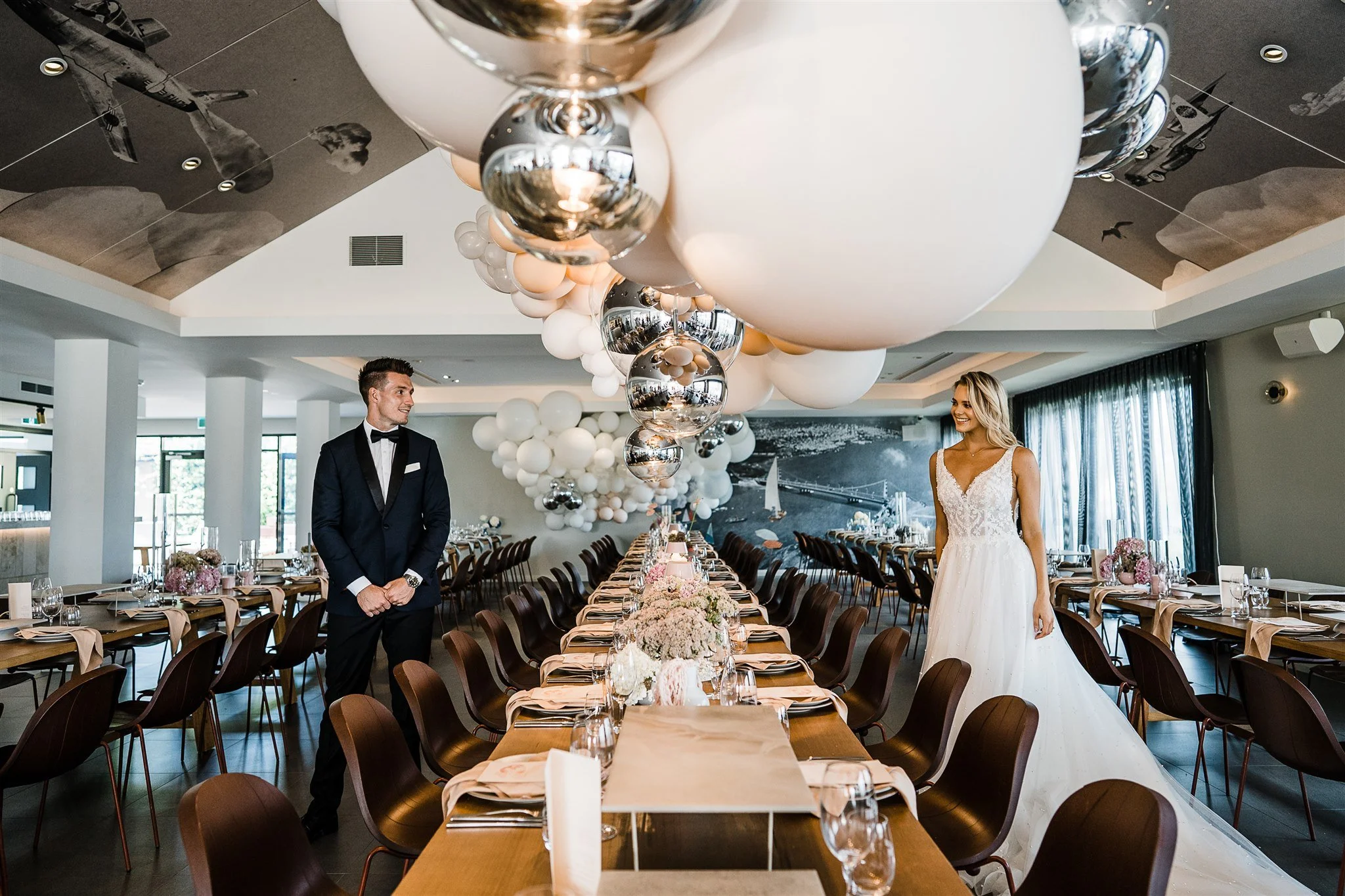 A wedding reception decorated with large white and silver balloons hanging from the ceiling, with a long table set with flowers, tableware, and napkins. A bride in a white wedding gown and a groom in a black tuxedo are standing and smiling in the background.