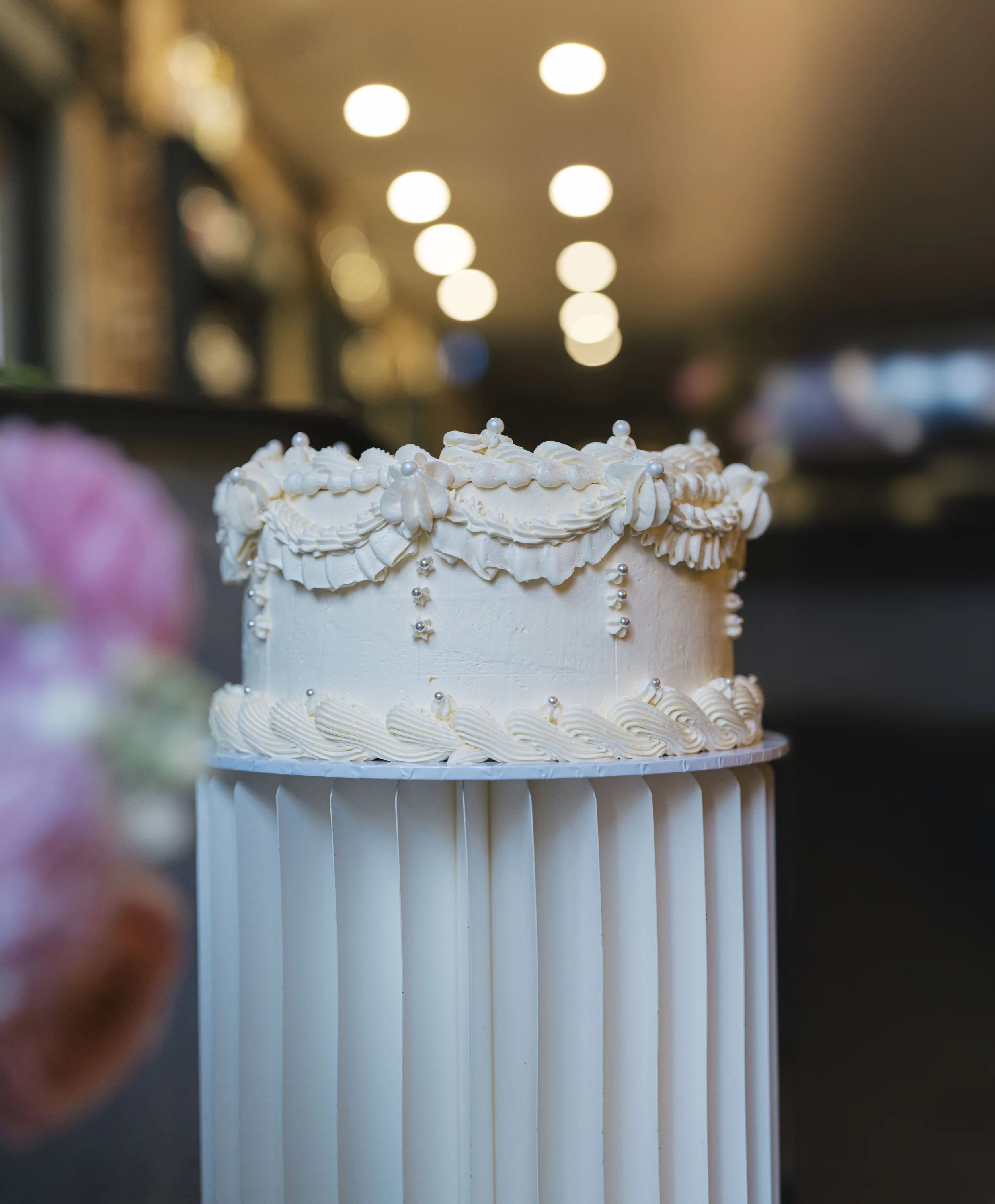 Close-up of a decorated white birthday cake with piped cream, pearl-like decorations, and silver dragees, with a blurred background of warm ceiling lights.