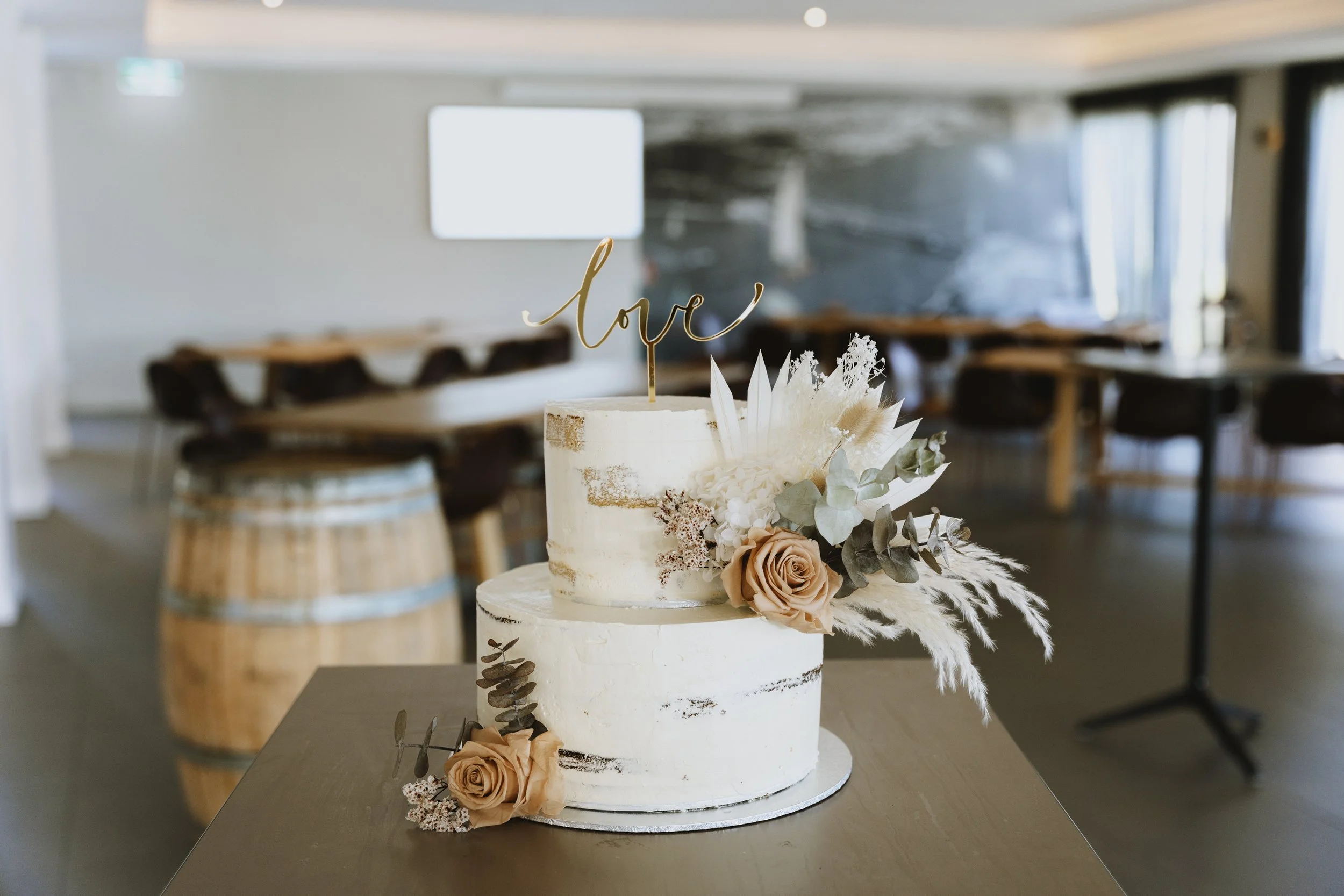A wedding cake decorated with beige roses, white flowers, and eucalyptus leaves, with a gold 'love' cake topper, displayed on a table in an event space.