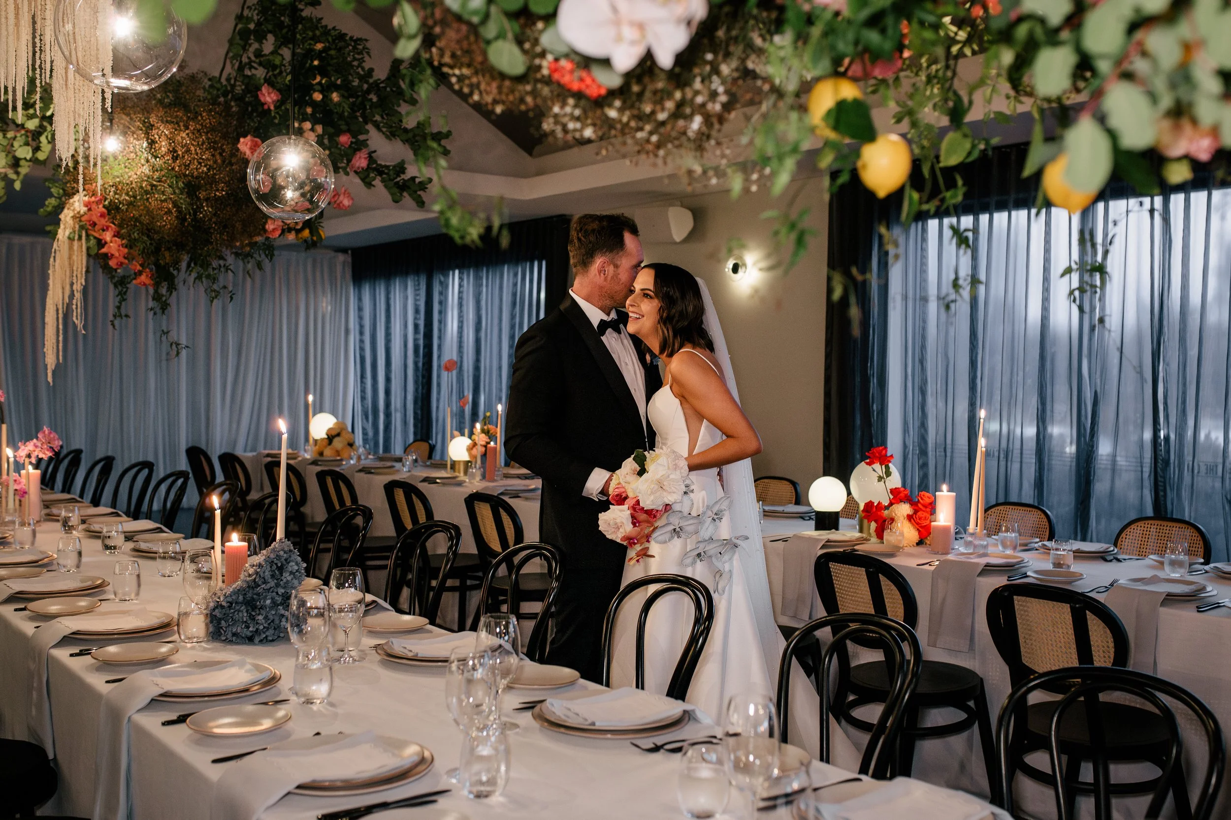 A bride and groom are standing close together inside a decorated wedding reception hall with tables set for dinner and floral arrangements hanging from the ceiling.