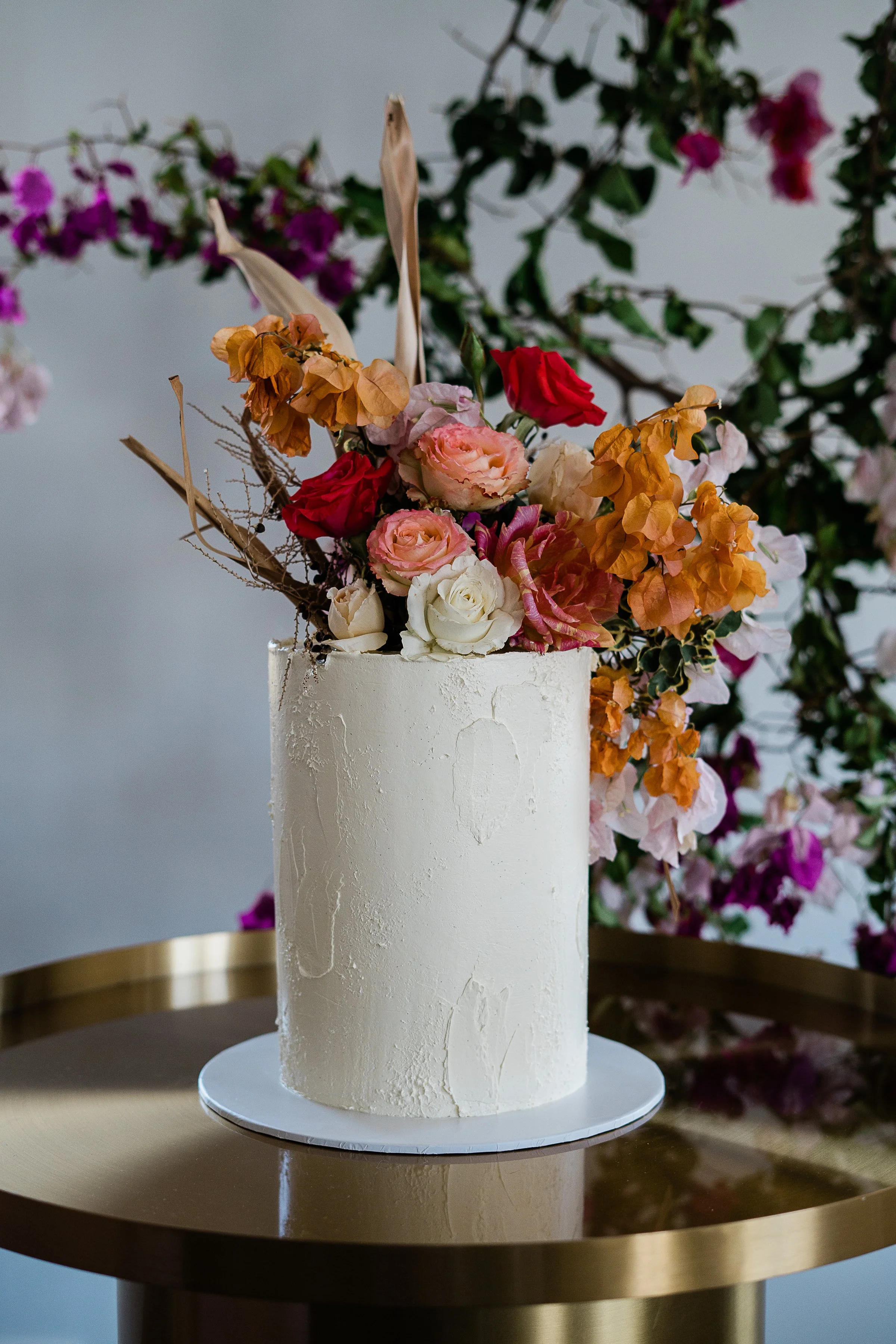 A tall white wedding cake with a smooth, textured finish, decorated with a cascading arrangement of assorted pink, red, orange, and white flowers, placed on a round white cake board on top of a gold table.