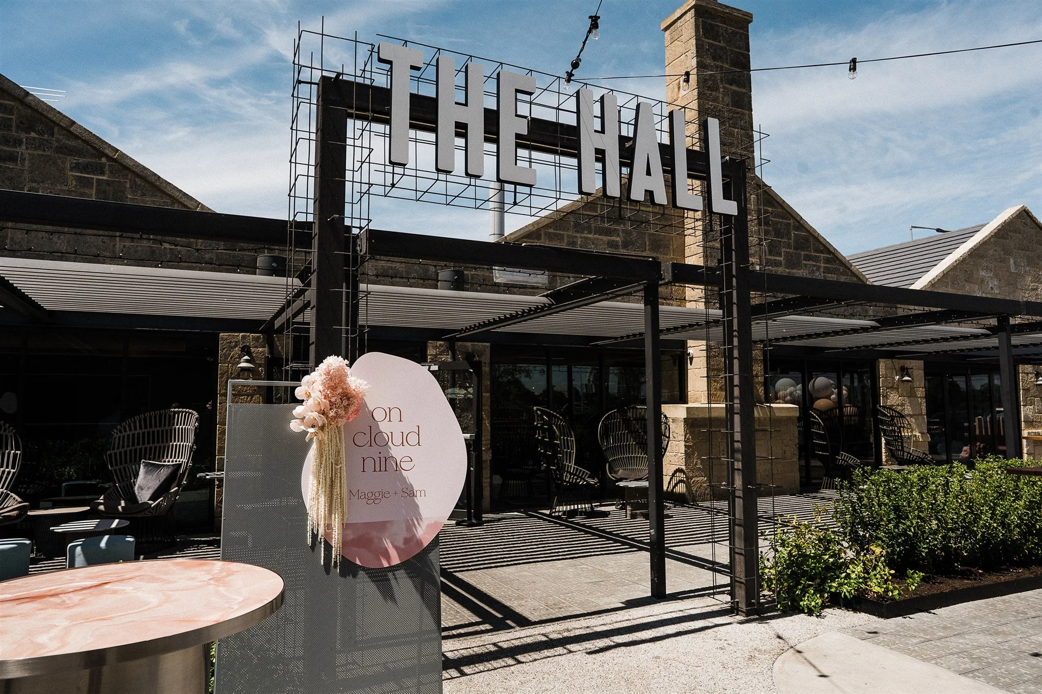 Exterior of a restaurant or event venue with a large sign that reads 'THE HALL' on a metal framework. There is outdoor seating with modern chairs and a pink and white sign with the words 'on cloud nine' and 'Maggie + Sam' on it. The building has stone walls and a chimney, with string lights hanging overhead. The sky is partly cloudy with blue visible.
