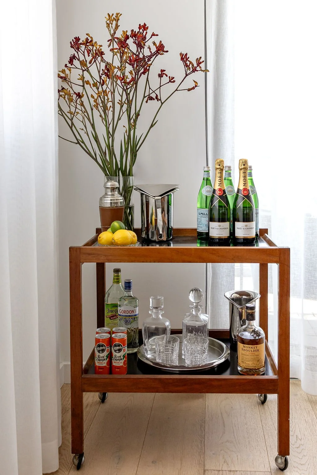 A mid-century style bar cart with two shelves. On the top is a vase with tall Kangaroo Paws in it, a small bowl with lemons and several bottles of champagne. On the lower shelf is a round silver tray with glass decanters, some bottles and cans of dri