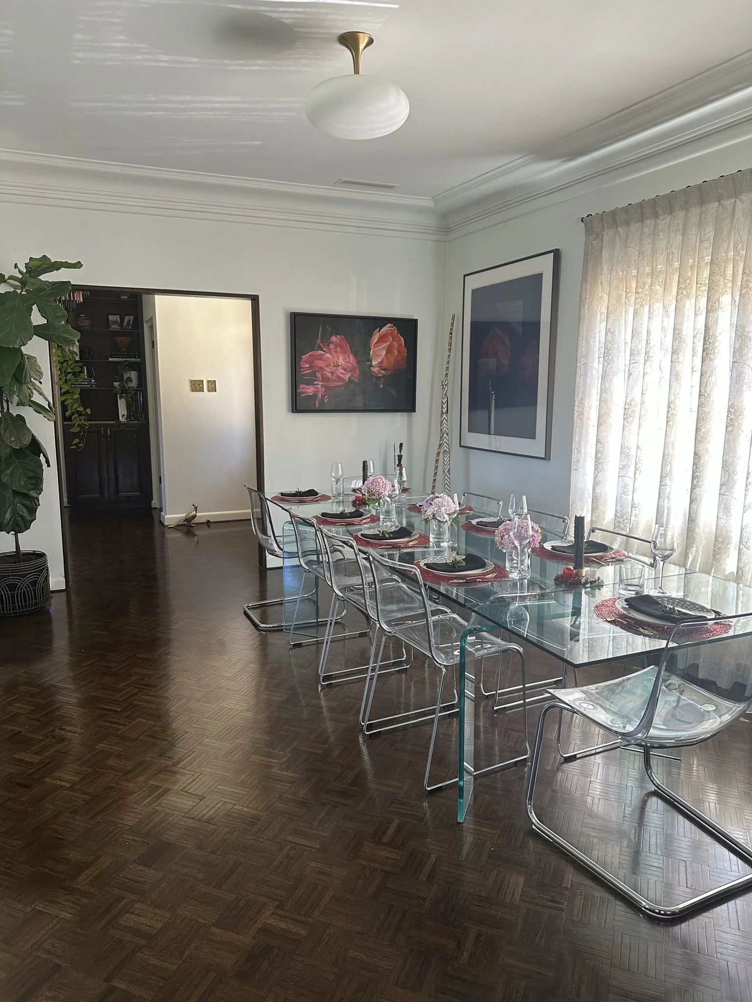 A long glass dining table and chairs is set for dinner. Behind it is a painting of flowers. The light fixture is elegant and the floors are dark stained parquet.