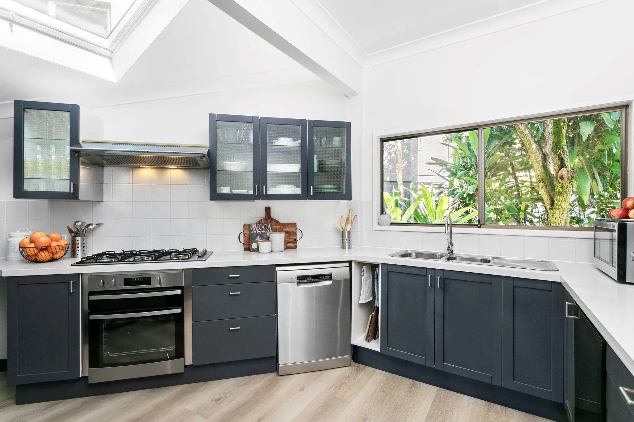 A kitchen in an L shape, with beautiful green trees outside the window splashback. The walls are white and the cabinets are dark blue. The space is modern and stylish.