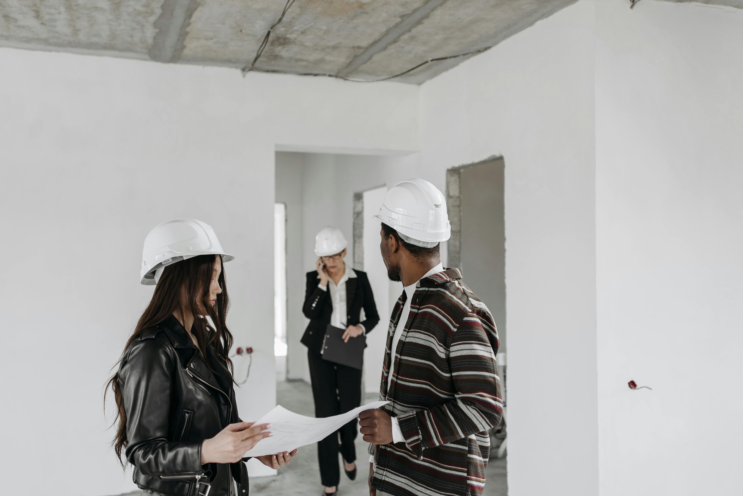 Three people wearing white hard hats, one holding a plan and one on the phone. Two are women, one is a man.