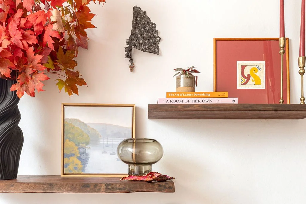 An image of a mantlepiece with bright autumn leaves in a vase, and artworks and books on the shelf.