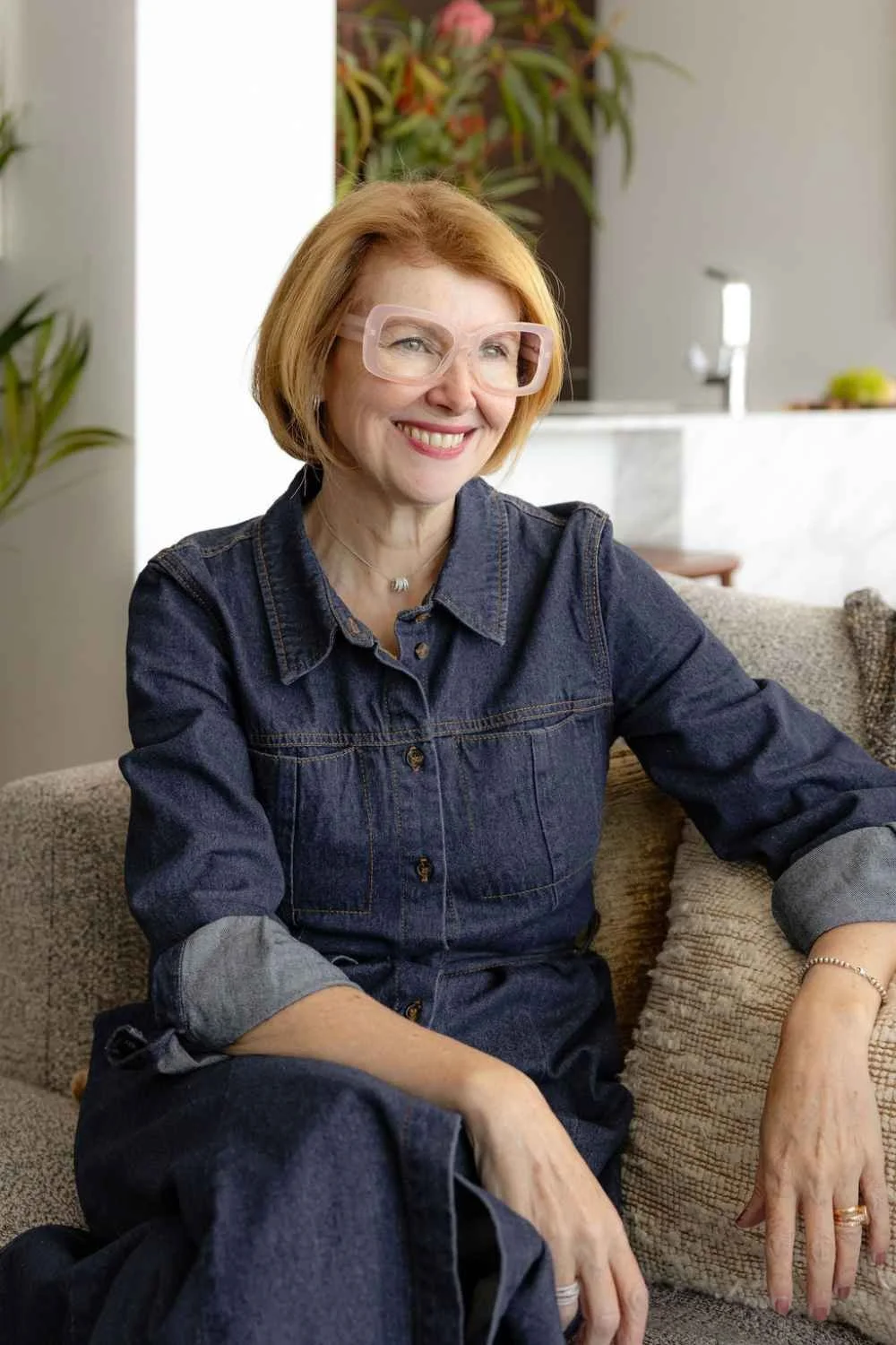 Kate Kerin seated indoors wearing a dark denim outfit and pale pink glasses, smiling in a softly styled living space.