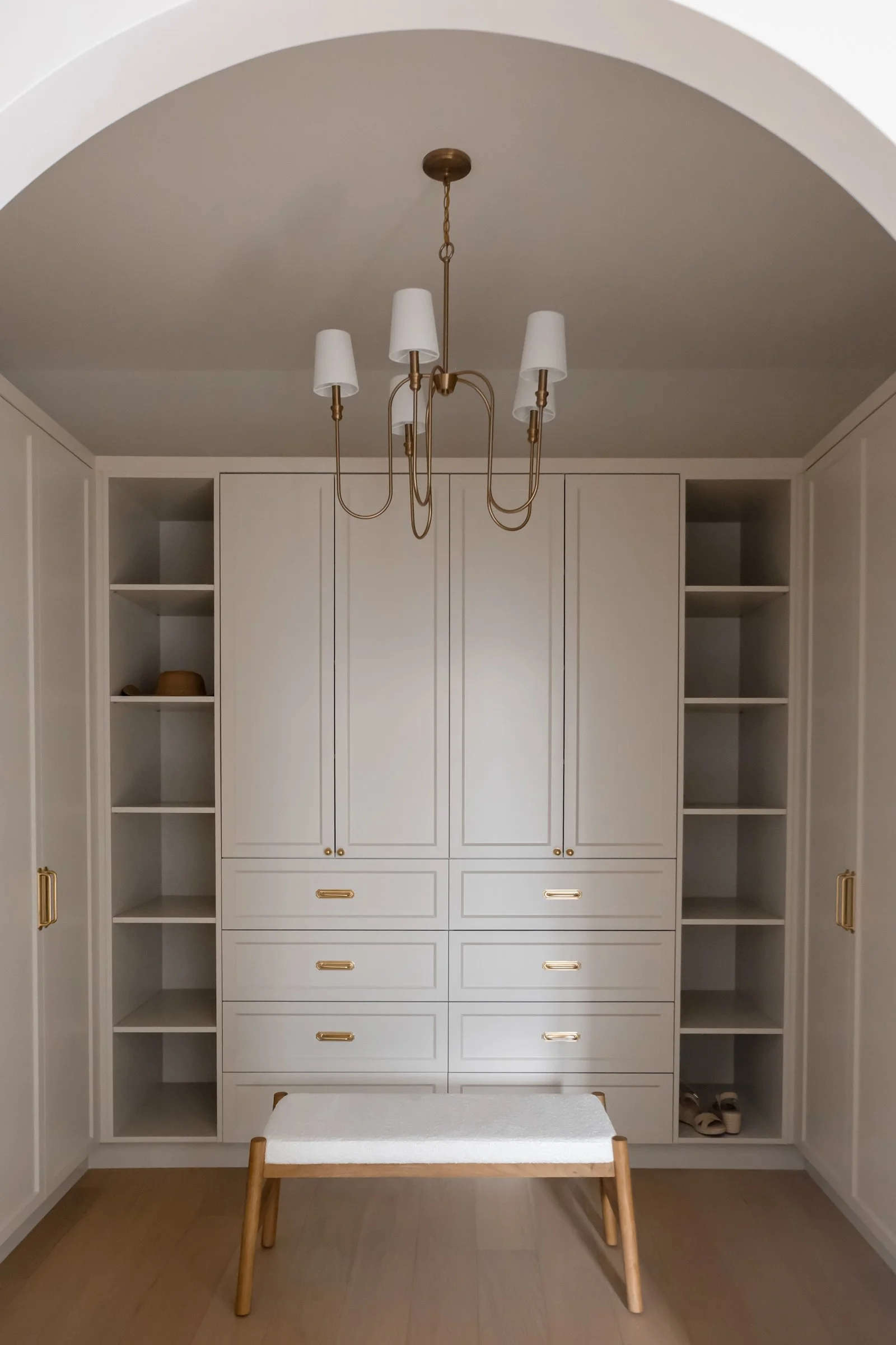 An elegant wardrobe with open shelves, and cupboards. In front is a feature pendant light and a bench for sitting on.