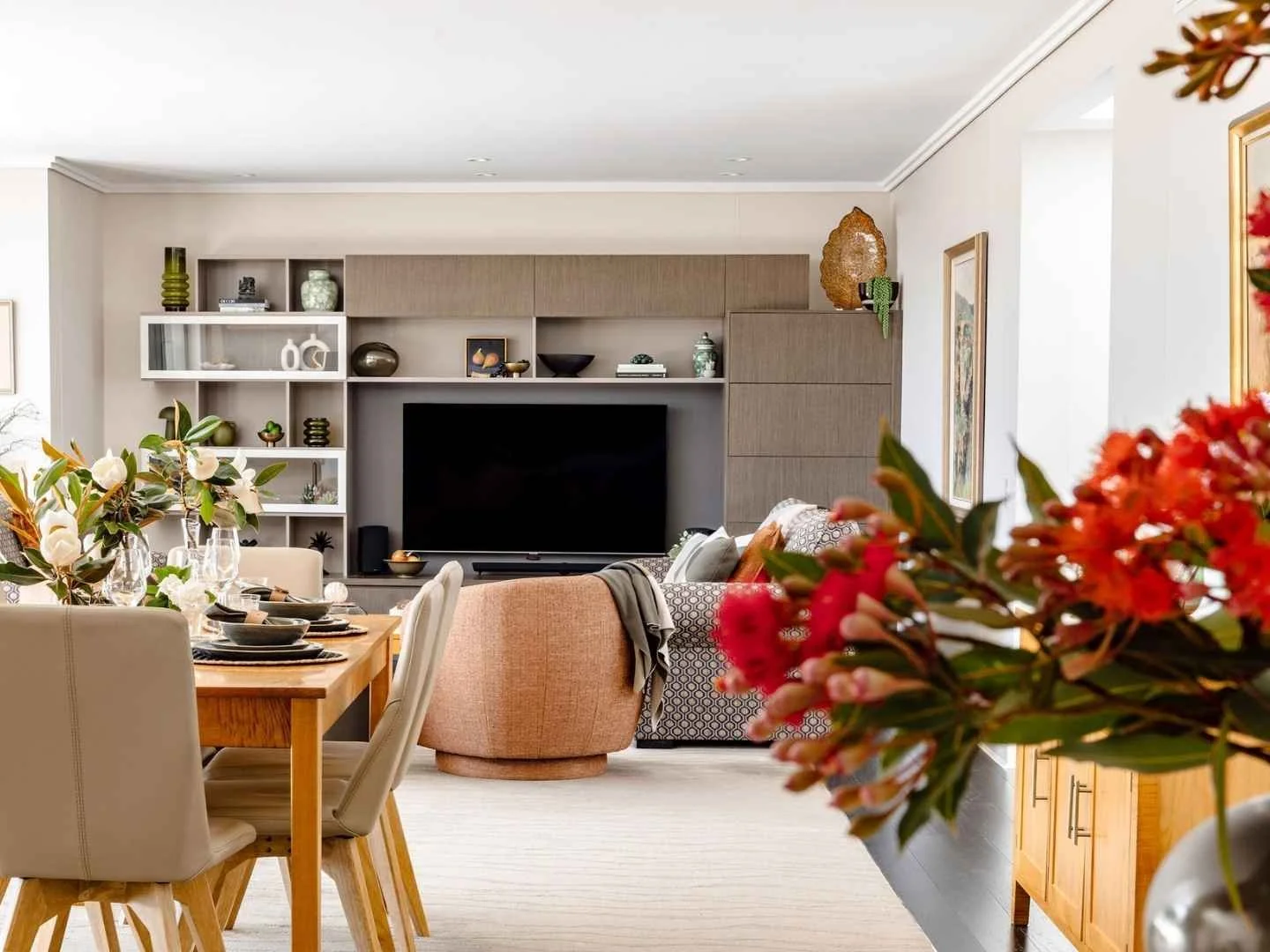 Open-plan living and dining interior with built-in cabinetry, dining table, curved armchair and red floral arrangement in the foreground.