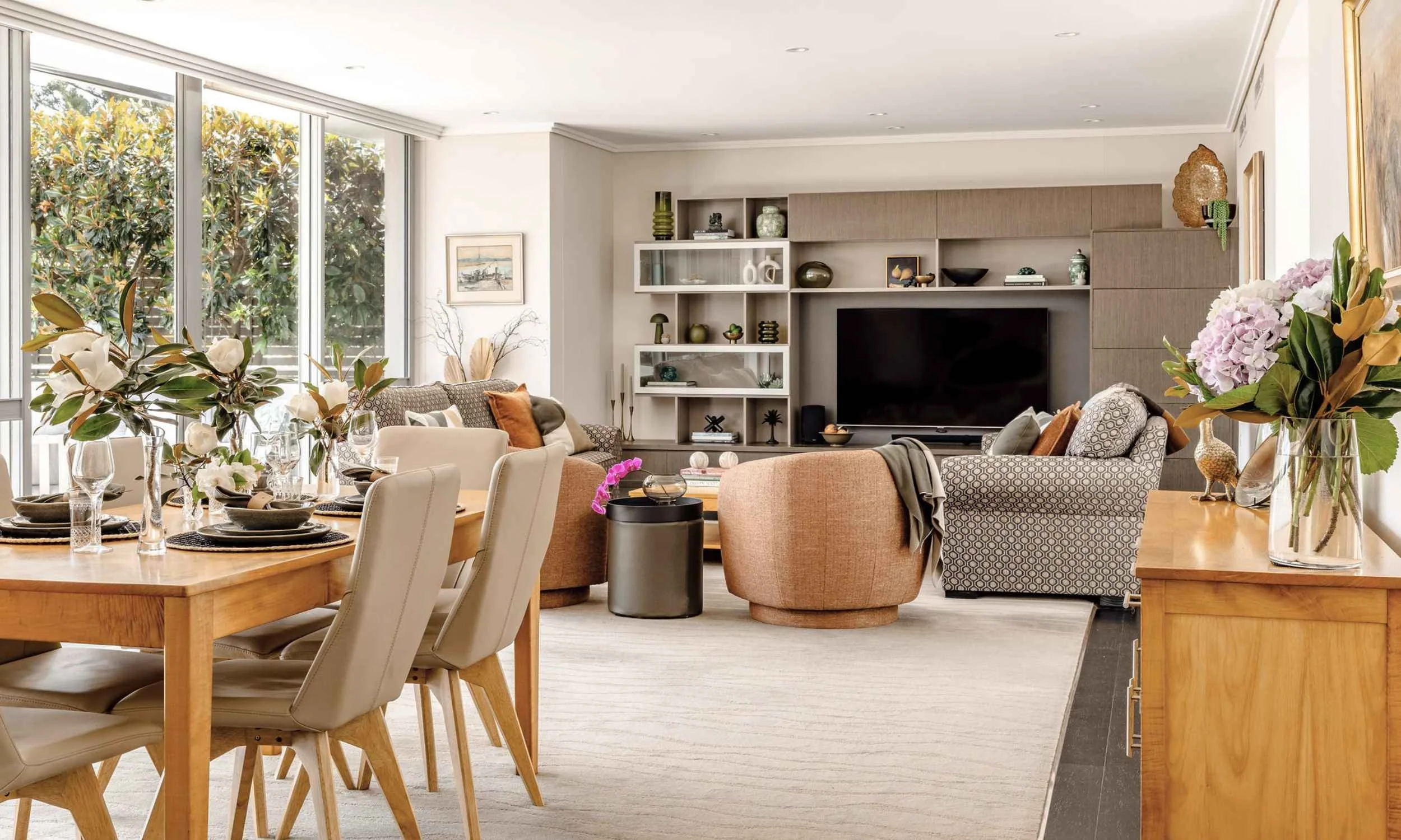 Open-plan Greenwich apartment interior with dining area, layered seating, built-in cabinetry and large windows overlooking greenery.