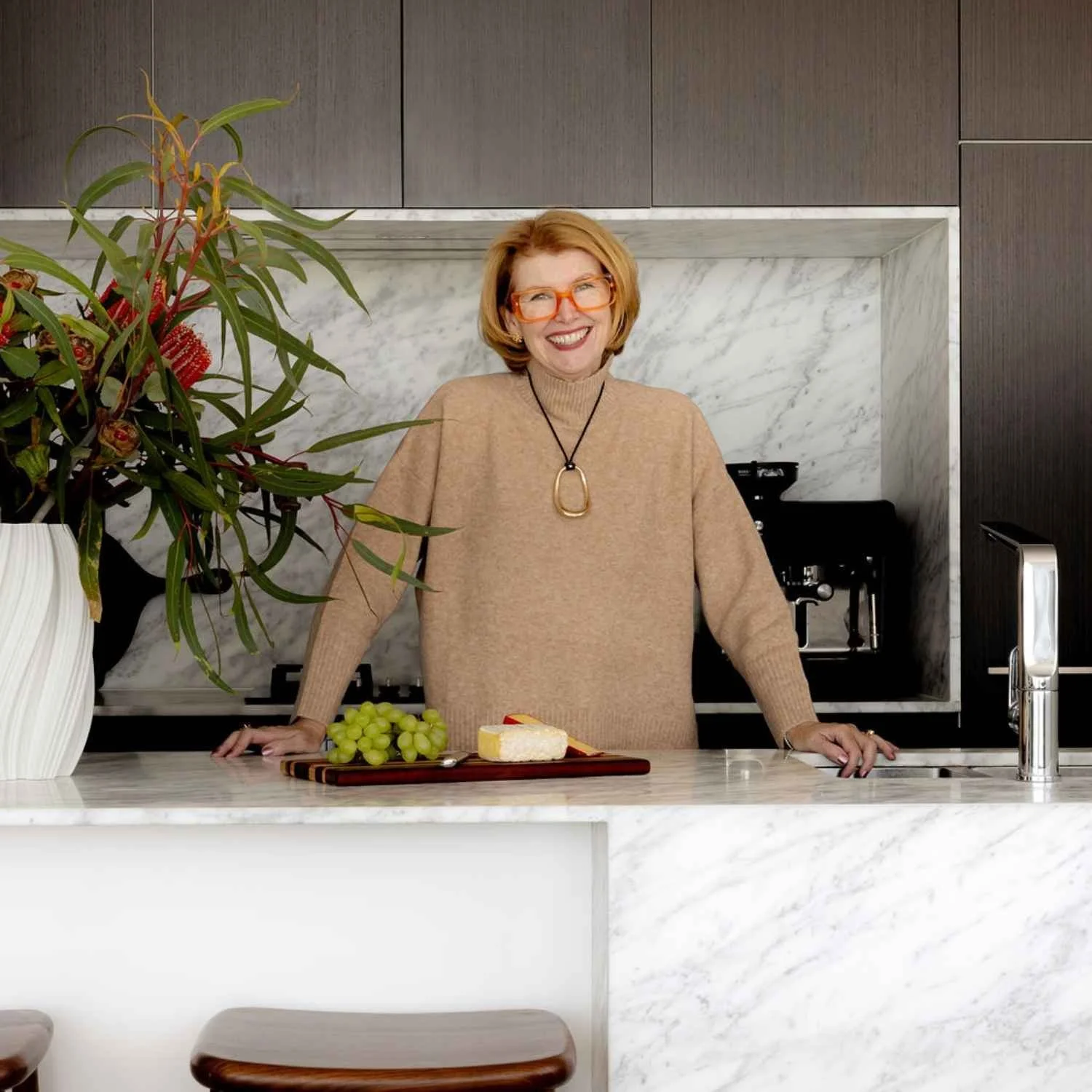 Kate standing behind a marble kitchen island in a contemporary North Sydney kitchen with dark cabinetry, cheese platter and a floral arrangement.