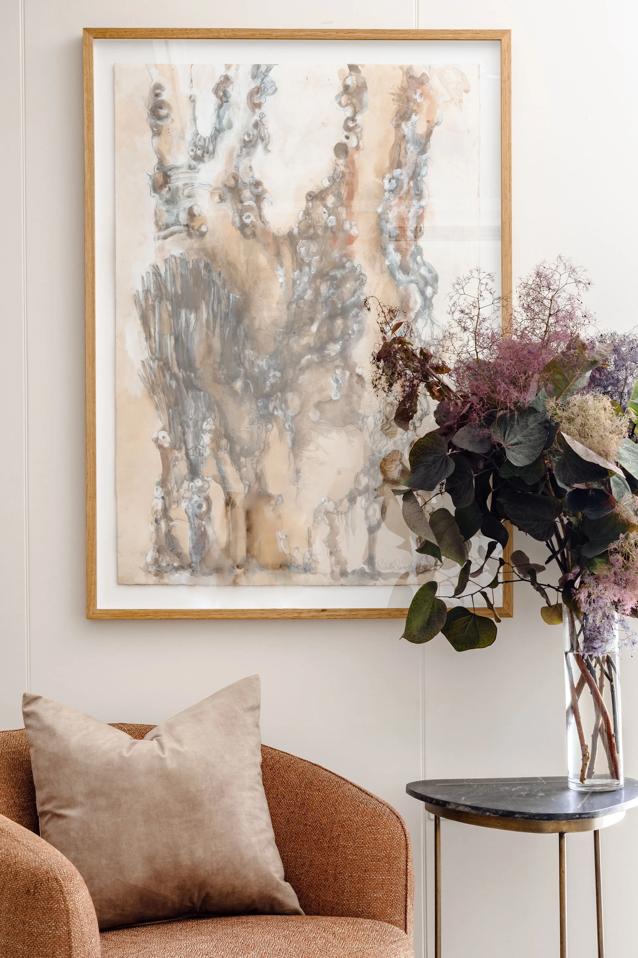 A beautiful painting, with a chair and soft cushion in front. A side table with flowers on it sits beside.